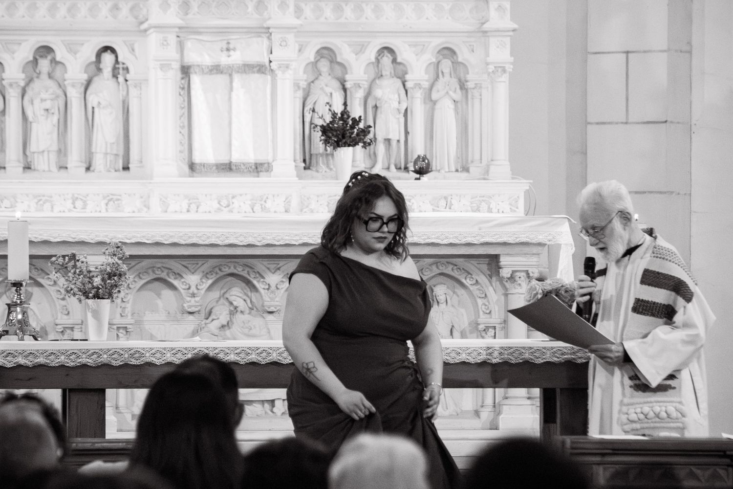 Black and white photo of a woman in a black dress receiving an award from a priest at a church altar ceremony.