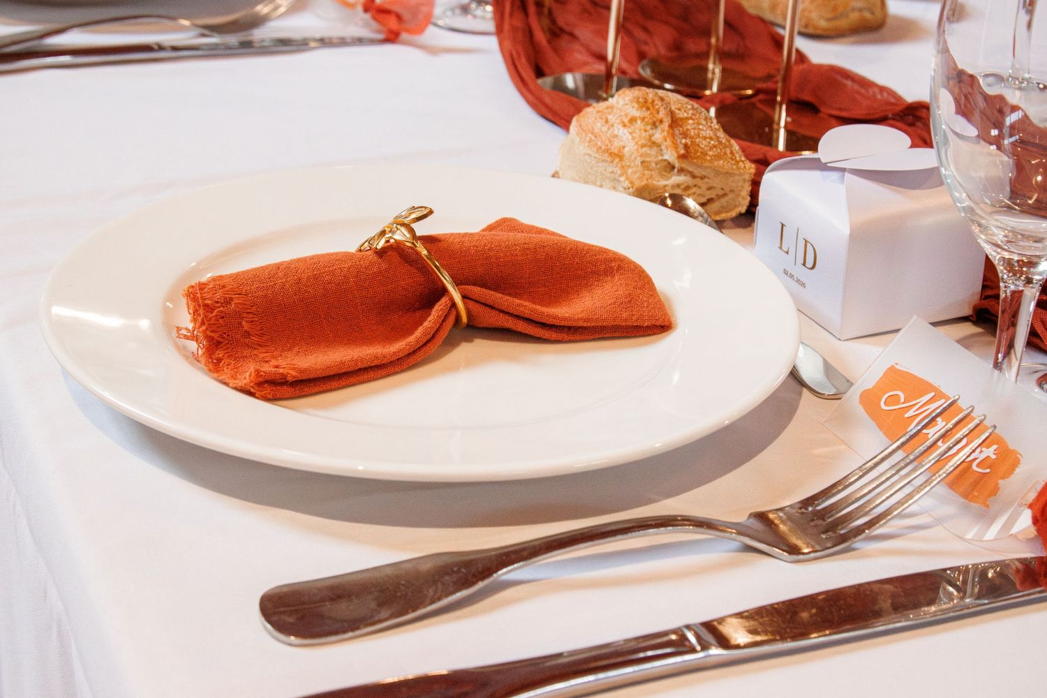 Elegant wedding table setting with terracotta folded napkin, white plate, silverware, and glassware on white tablecloth.