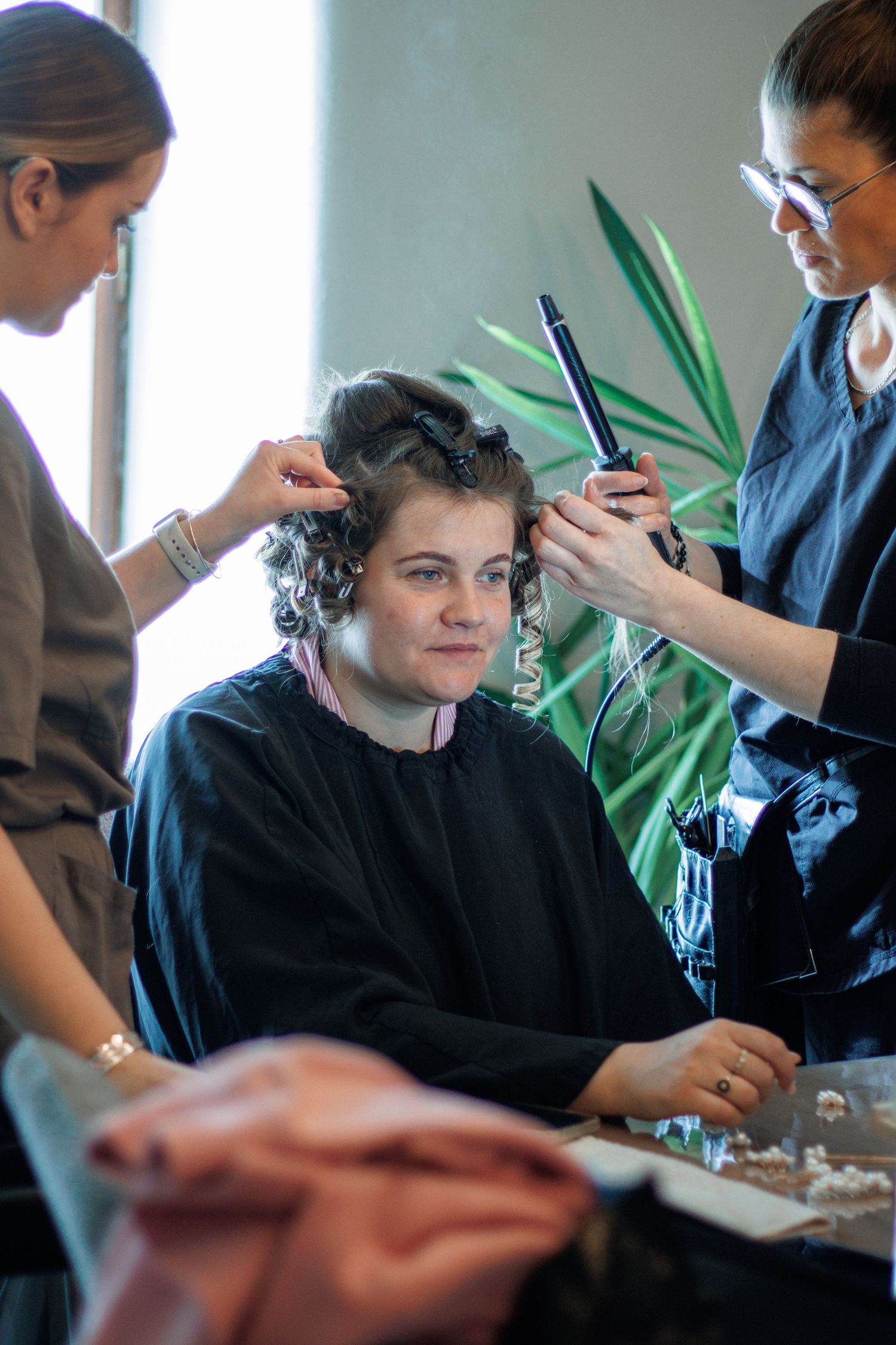 Two hairstylists working on a seated woman with curlers in a professional salon setting.