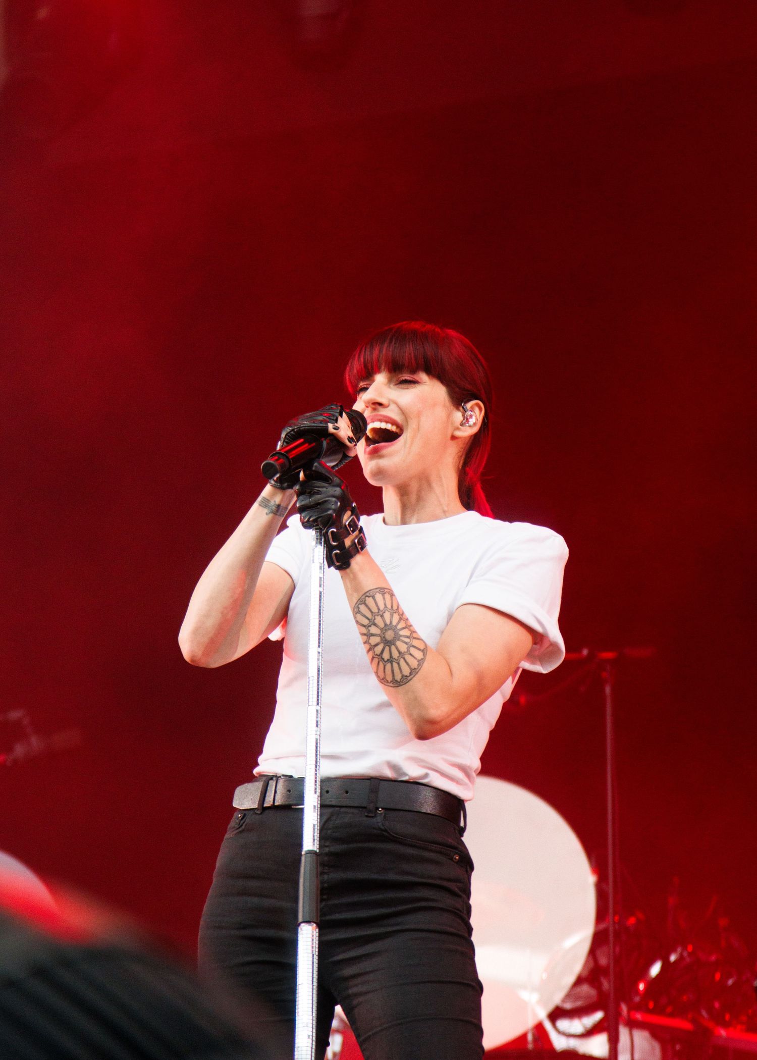 Female singer performing on stage, wearing white tee and black gloves, with red hair and arm tattoo.