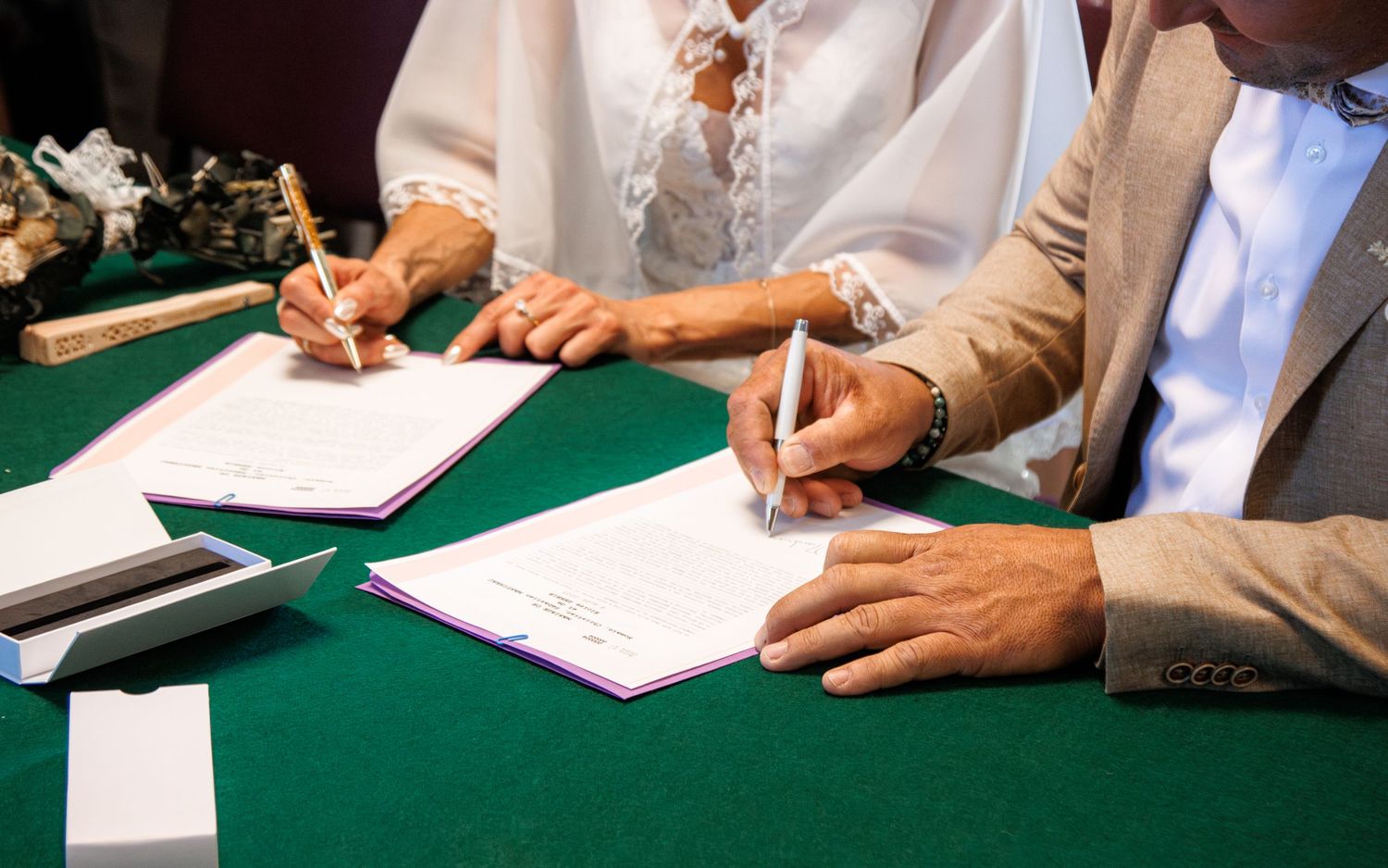 People in formal attire signing documents on a green table surface during a business meeting or ceremony.
