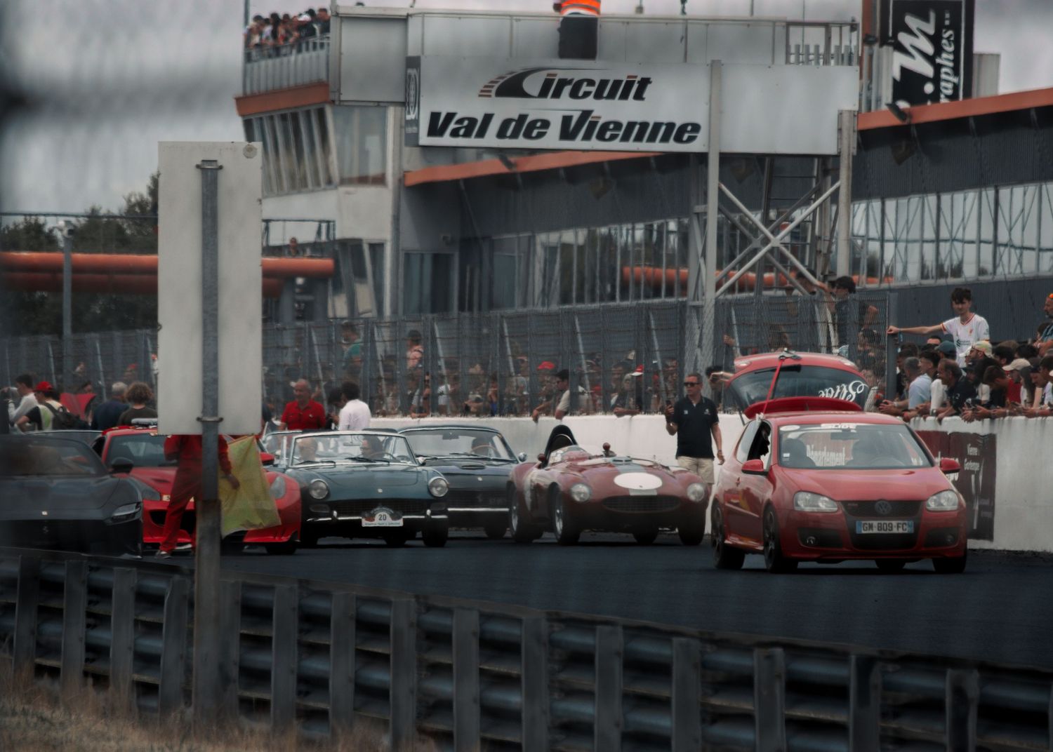 Classic cars racing at Val de Vienne circuit with pit lane structures visible in background.