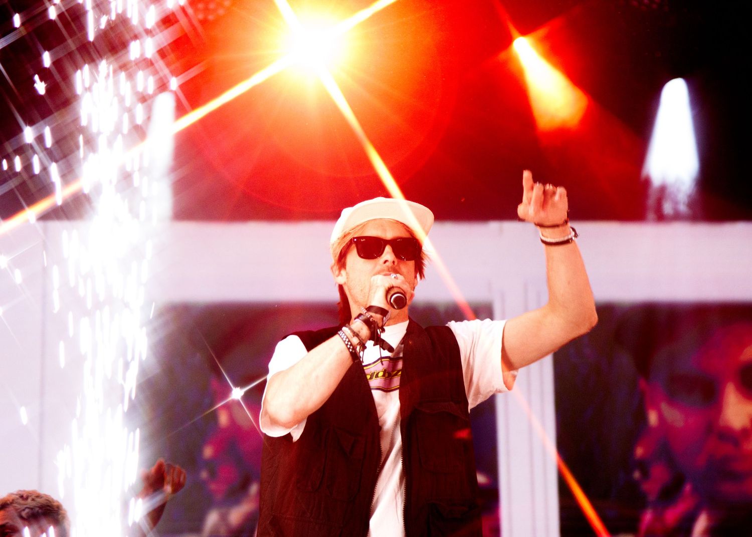 A performer in sunglasses and white cap sings into a microphone with dramatic red stage lighting in the background.