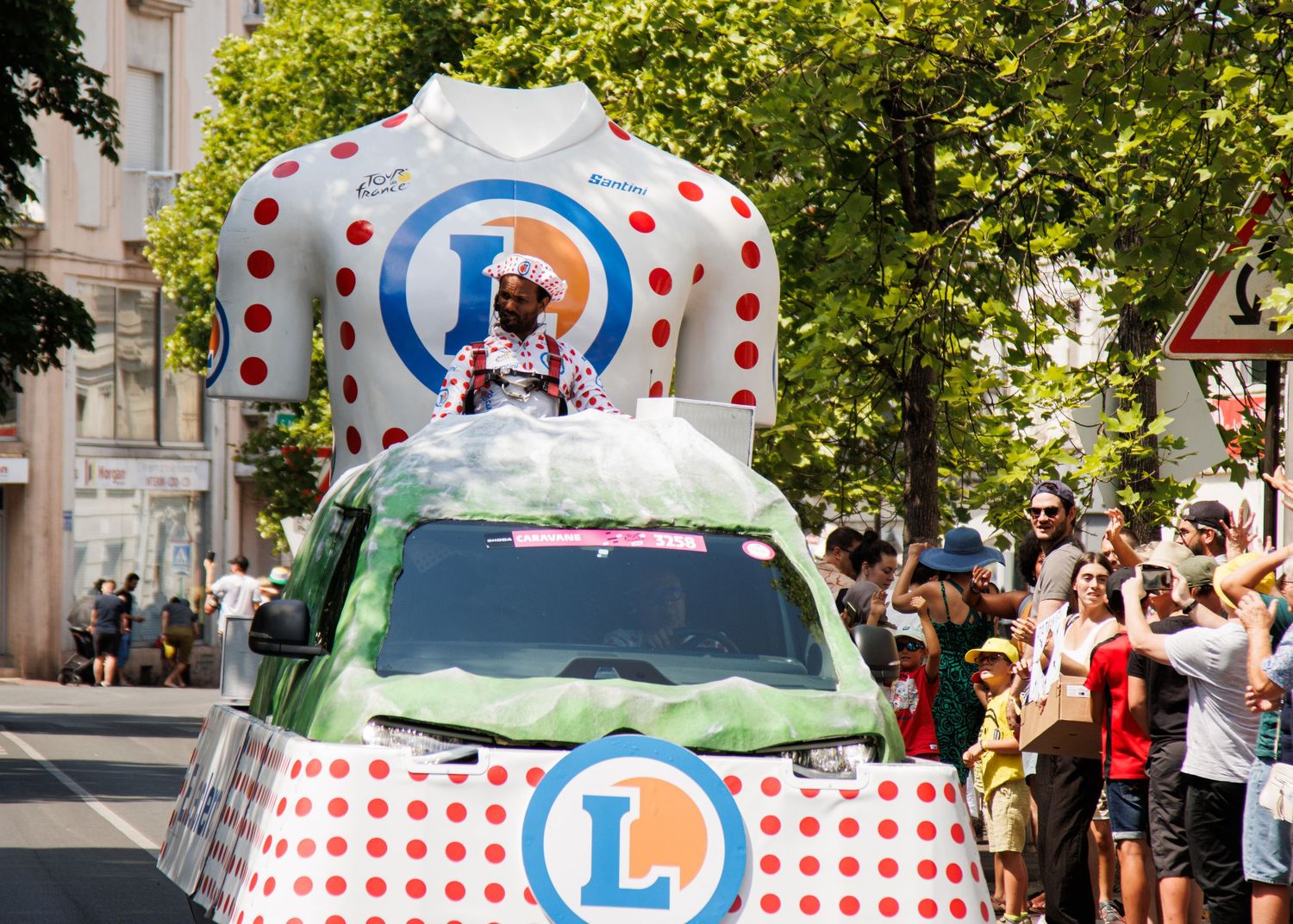 A promotional car with a polka-dotted design and large logo decoration drives in a parade while spectators watch.