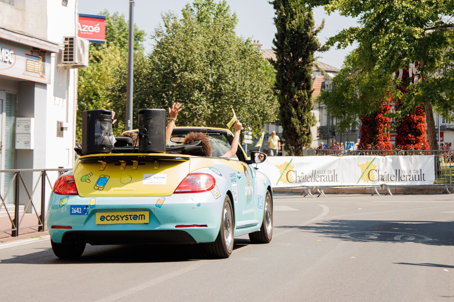 Cars and spectators line both sides of a tree-shaded urban street during a summer event.