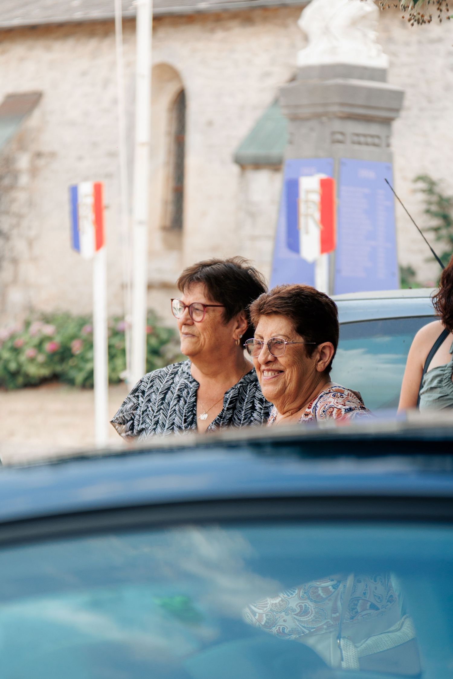 Two elderly women wearing glasses smile near French flags at an outdoor ceremony.