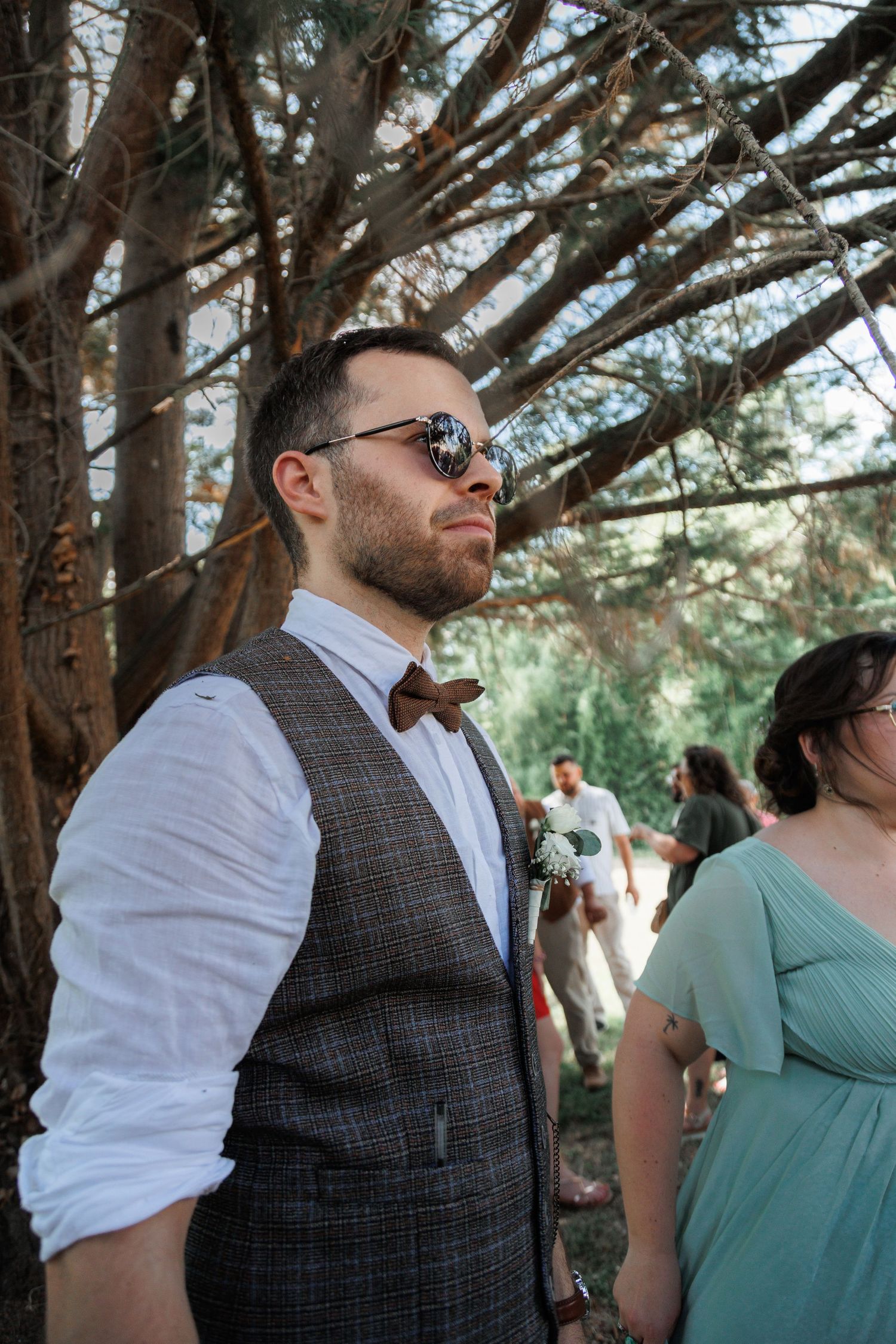 Stylish wedding guest wearing sunglasses and a brown bow tie vest stands under pine trees.