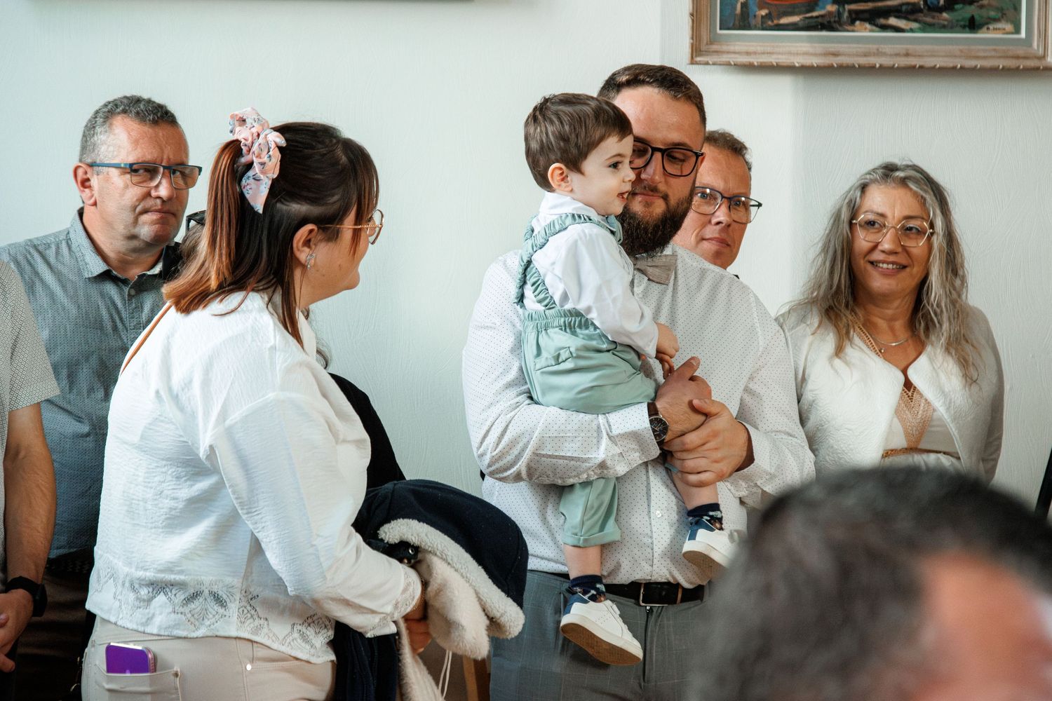 Family gathering indoors with man holding toddler, surrounded by smiling relatives in white attire at a celebration.