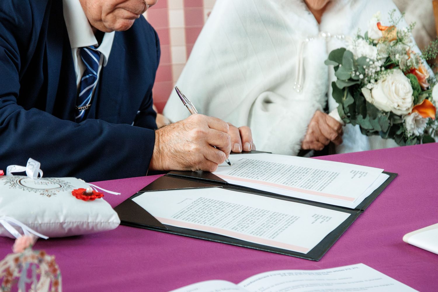 Elderly couple signing wedding documents at a ceremony table with bridal bouquet and ring pillow.