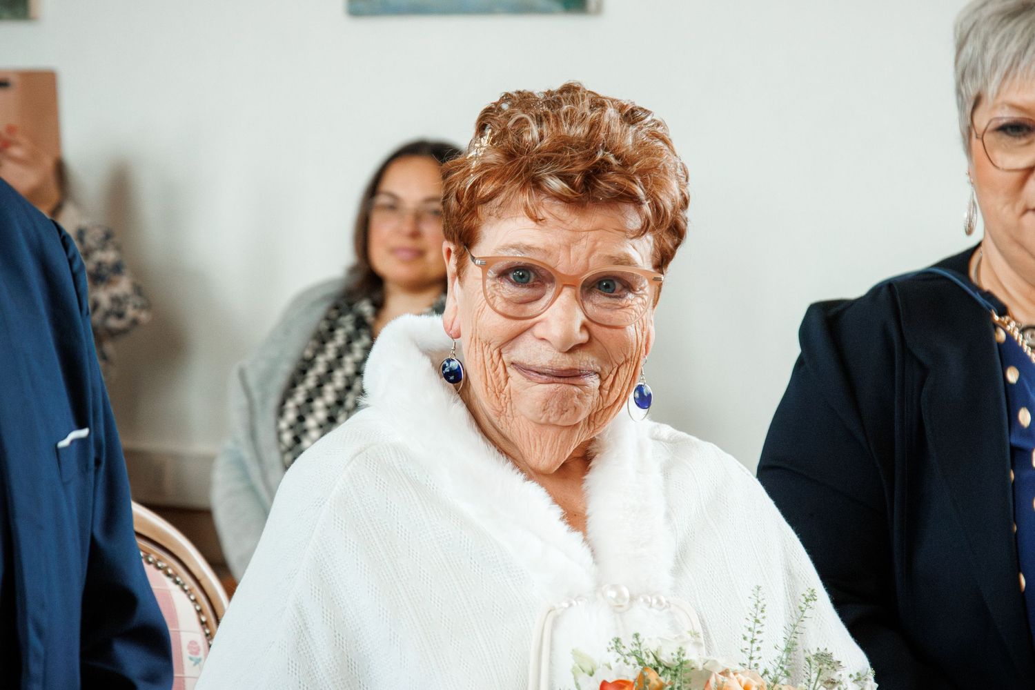 Elderly woman in white fur wrap smiling at a wedding ceremony, wearing blue earrings and holding flowers.