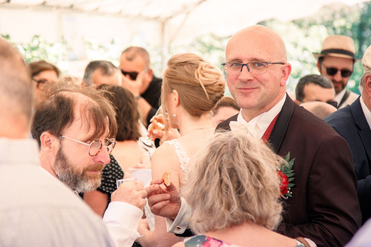 Bald groom in dark suit mingles with guests at outdoor wedding reception.
