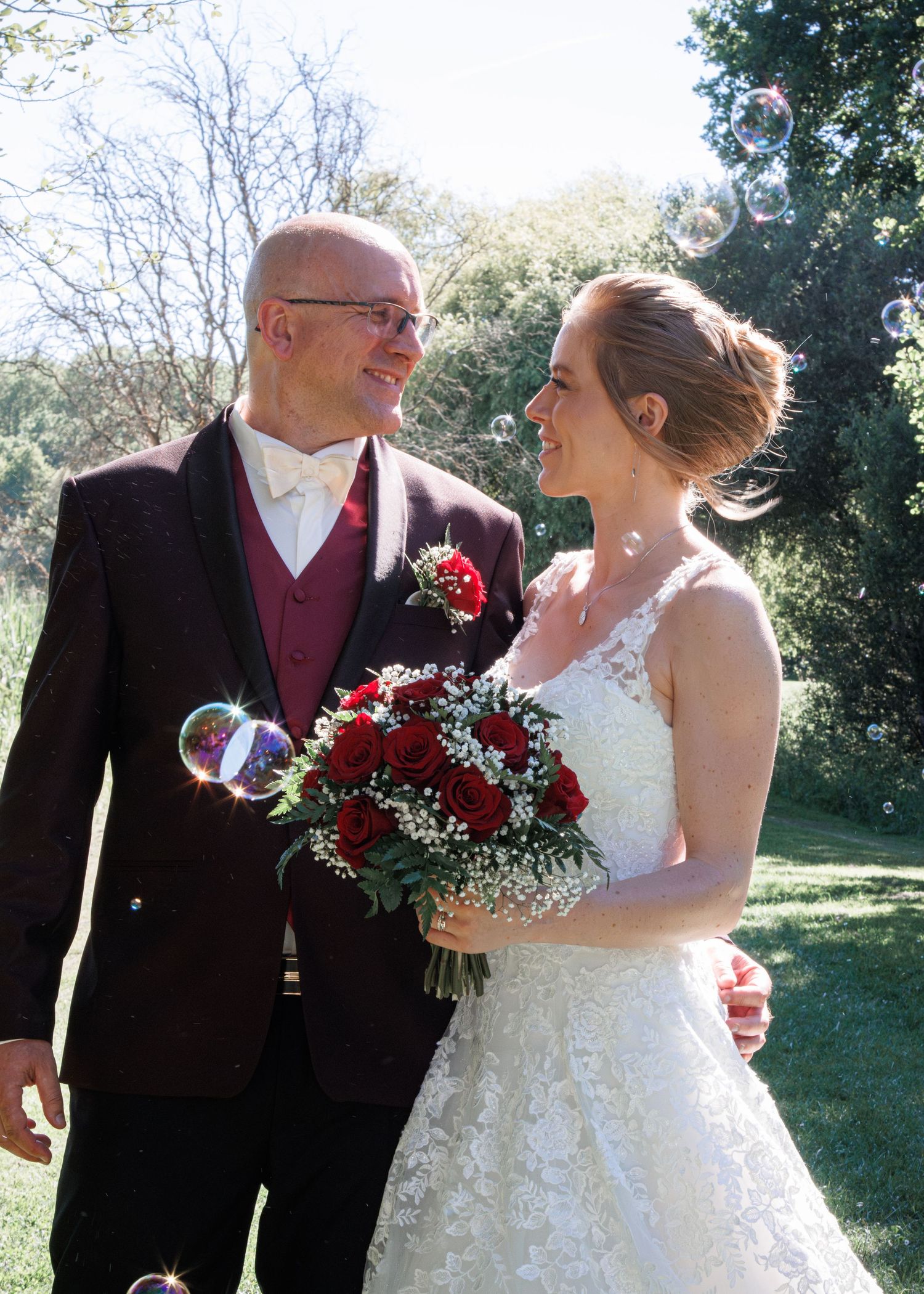 Happy bride and groom smiling at each other outdoors surrounded by soap bubbles, bride holding red rose bouquet in white lace dress.