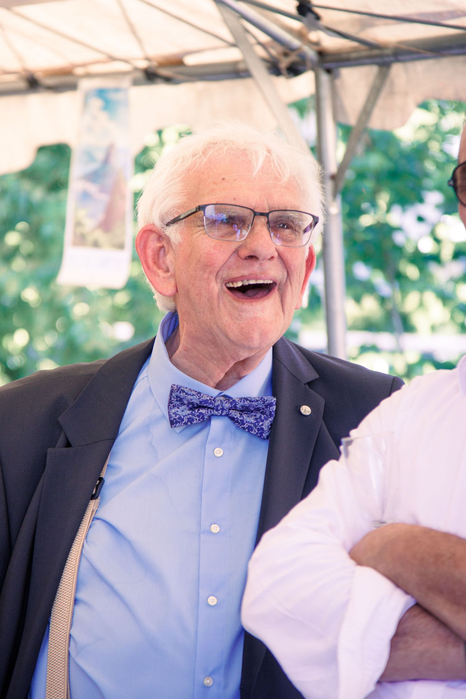 Elderly man in blue bow tie and blazer smiling and laughing at outdoor wedding celebration.