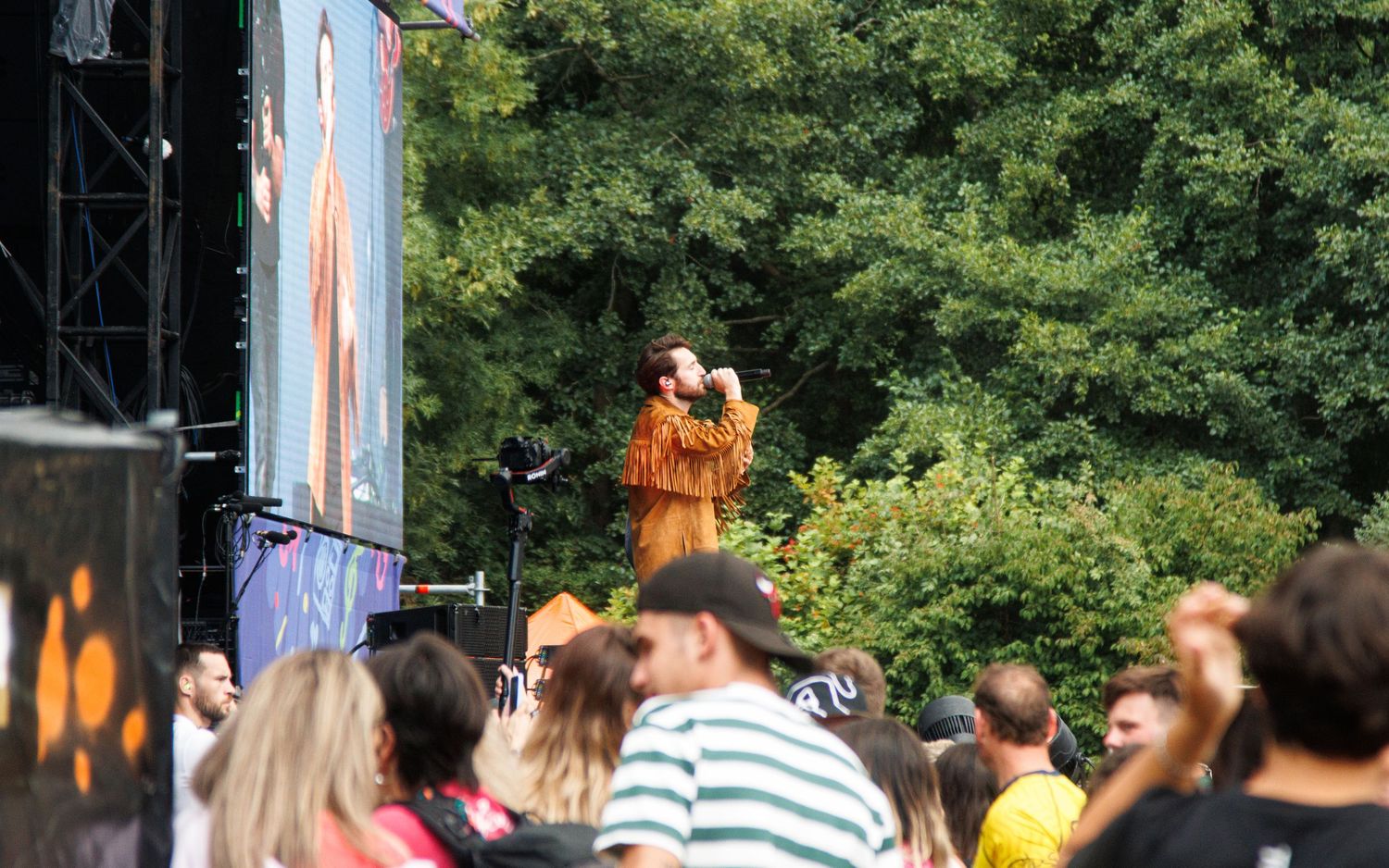 Crowd of people gathered outdoors watching a performance at a summer festival or event in a park setting.