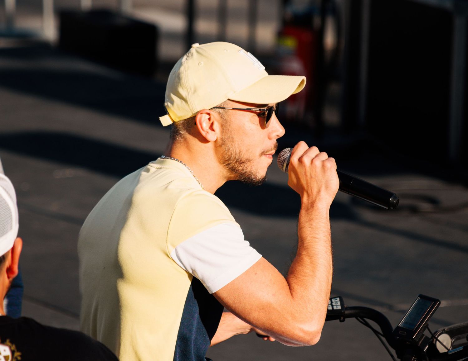 Performer in baseball cap and yellow shirt holds microphone while singing outdoors.