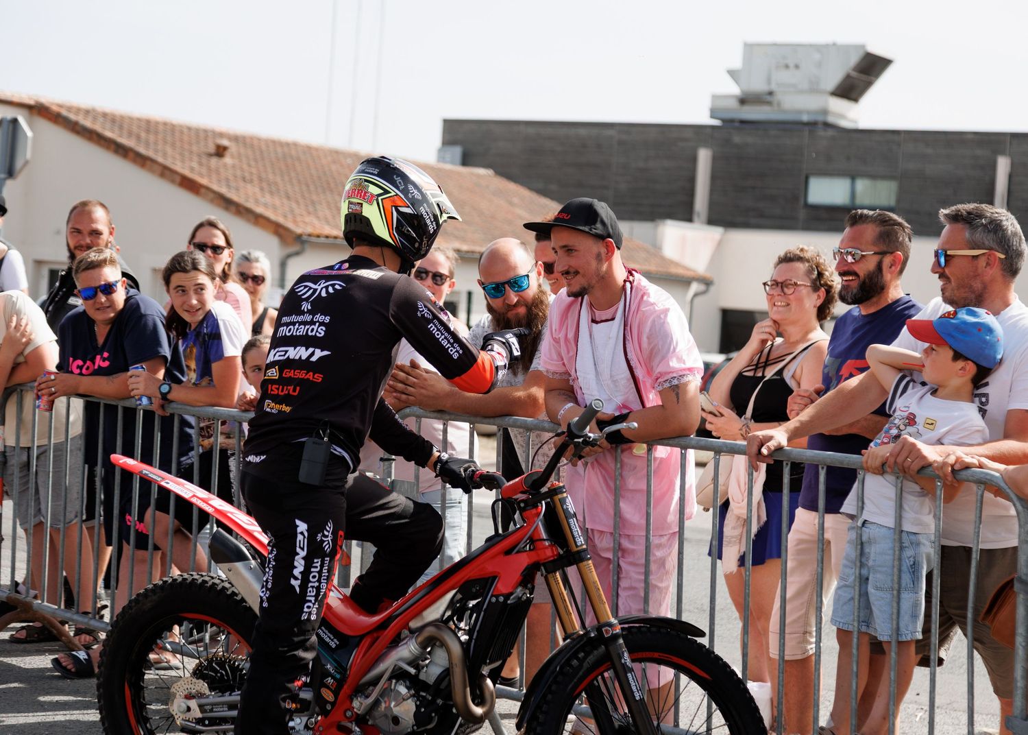 A motocross rider on a red bike greets fans at a sunny outdoor event, shaking hands over a metal barrier.