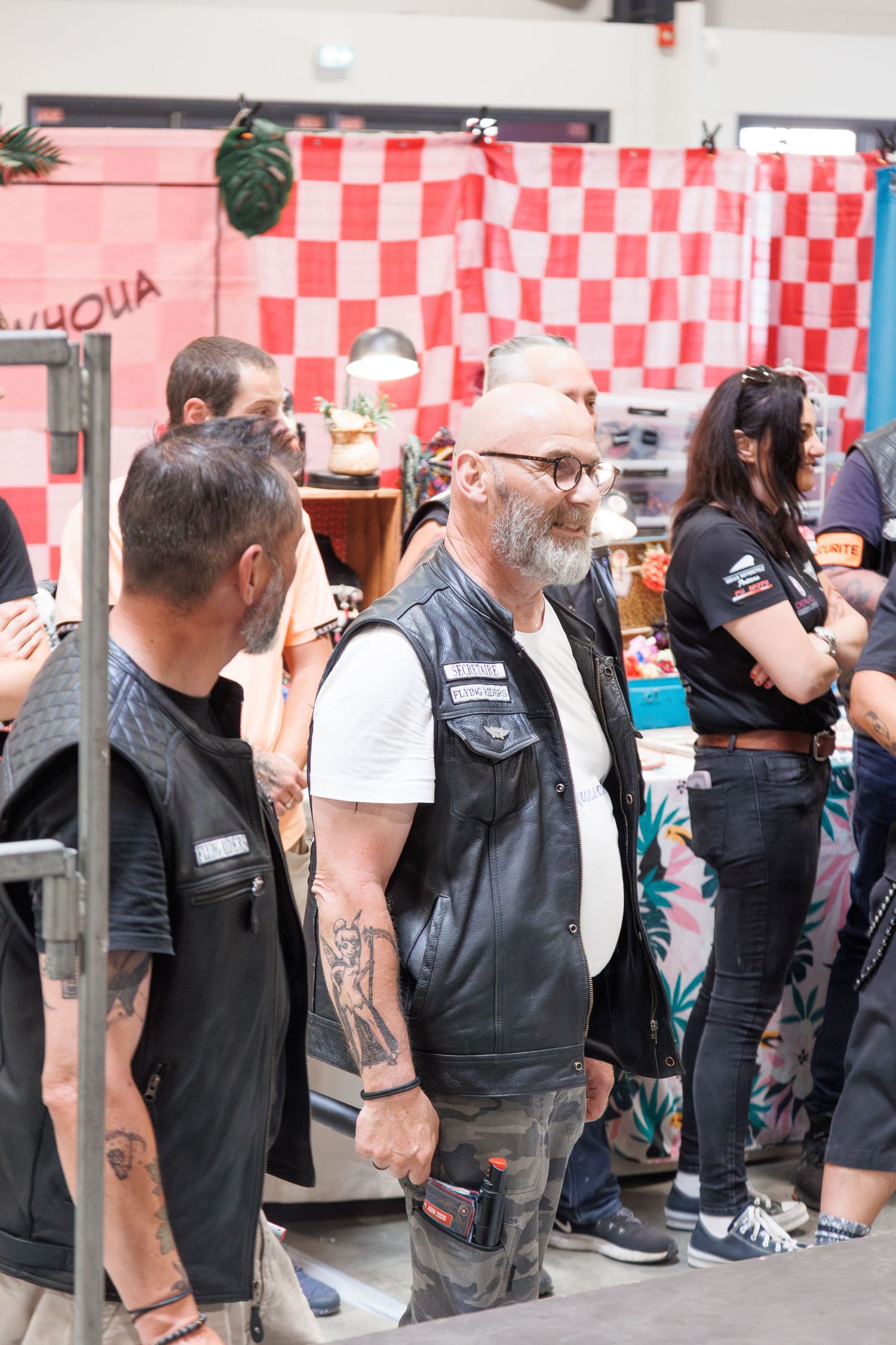Tattooed bikers in leather vests socialize at an indoor event with a red checkered backdrop.