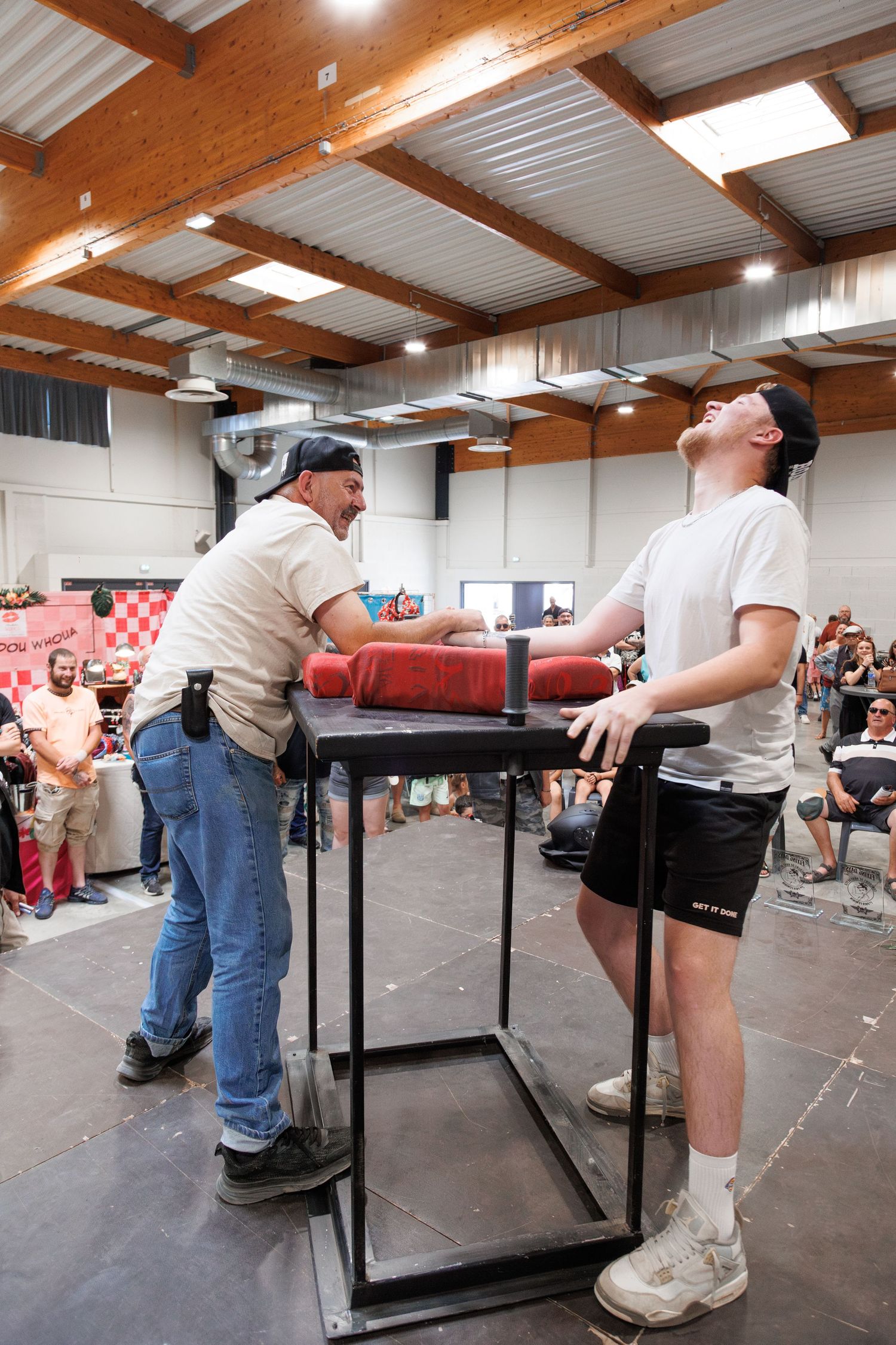 Two men compete in arm wrestling at an indoor sports event, with an audience watching in the background.