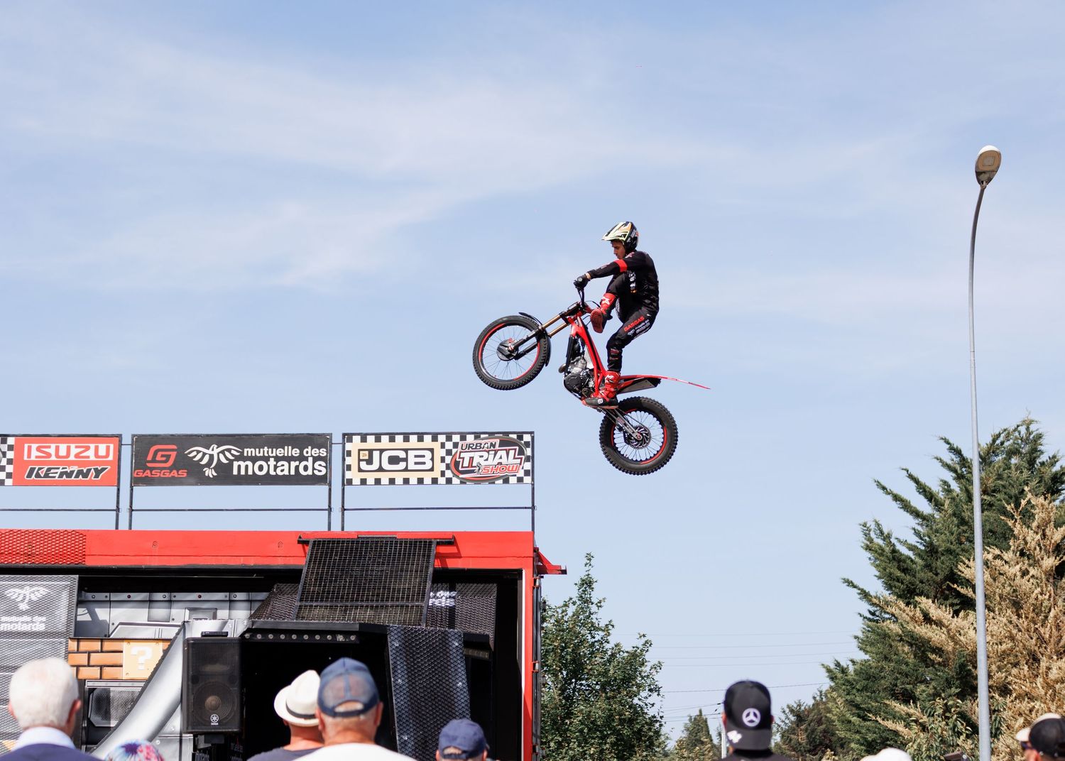 Motocross rider performing a dramatic wheelie jump stunt at an outdoor freestyle motorcycle show with crowd watching.
