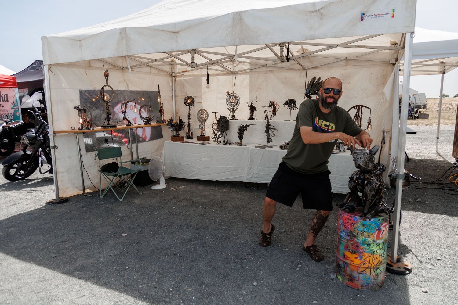 Man posing at outdoor art vendor booth displaying sculptures under white canopy tent at sunny open-air market.