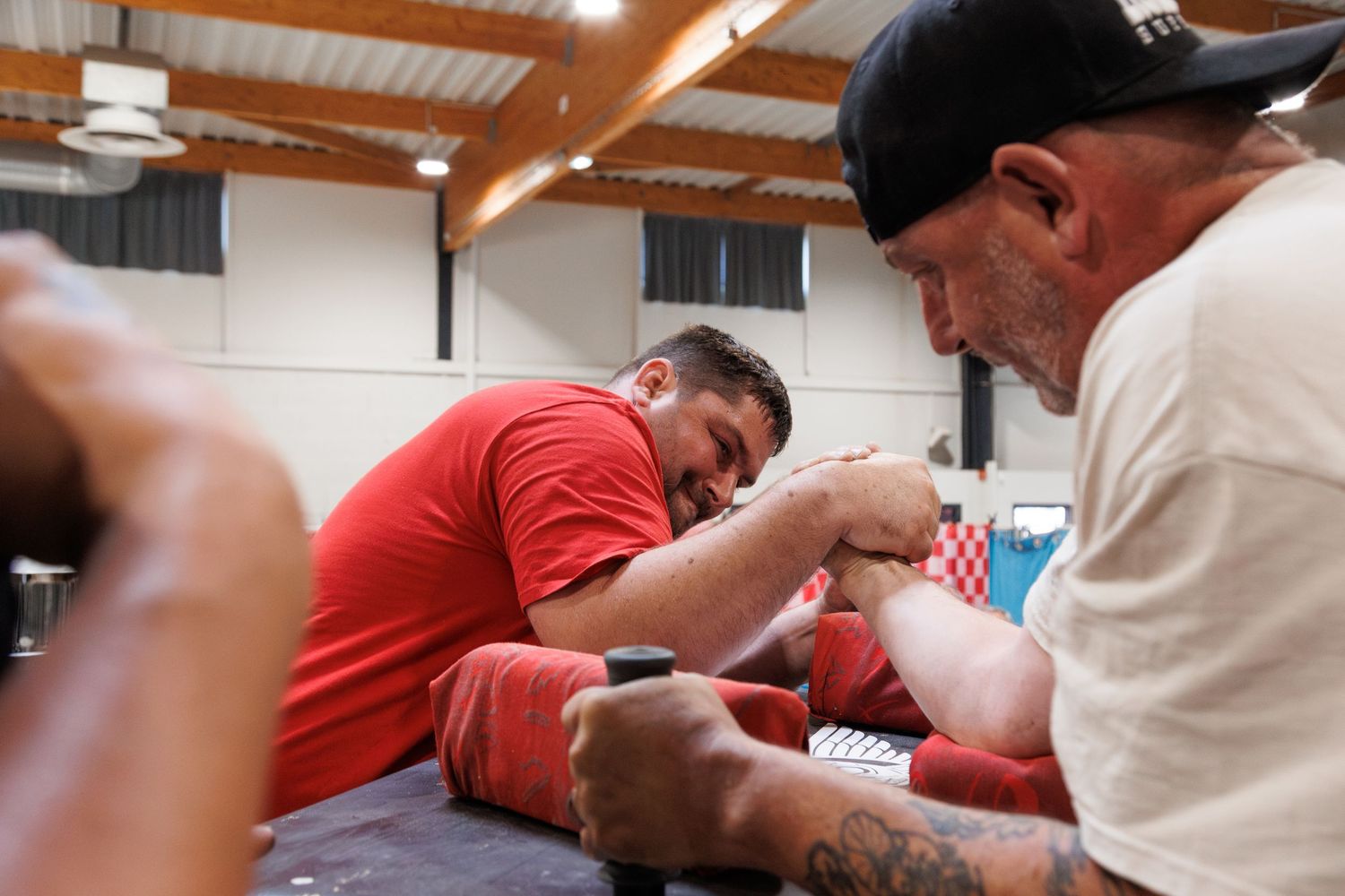 Two men competing in an arm wrestling match at an indoor tournament, one wearing a red shirt.