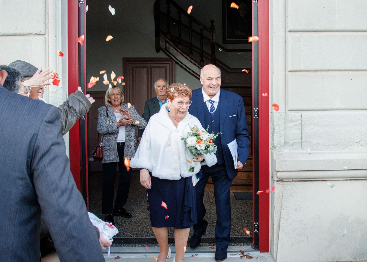 Elderly couple exits building as wedding guests throw confetti, bride holds bouquet in joyful outdoor ceremony.
