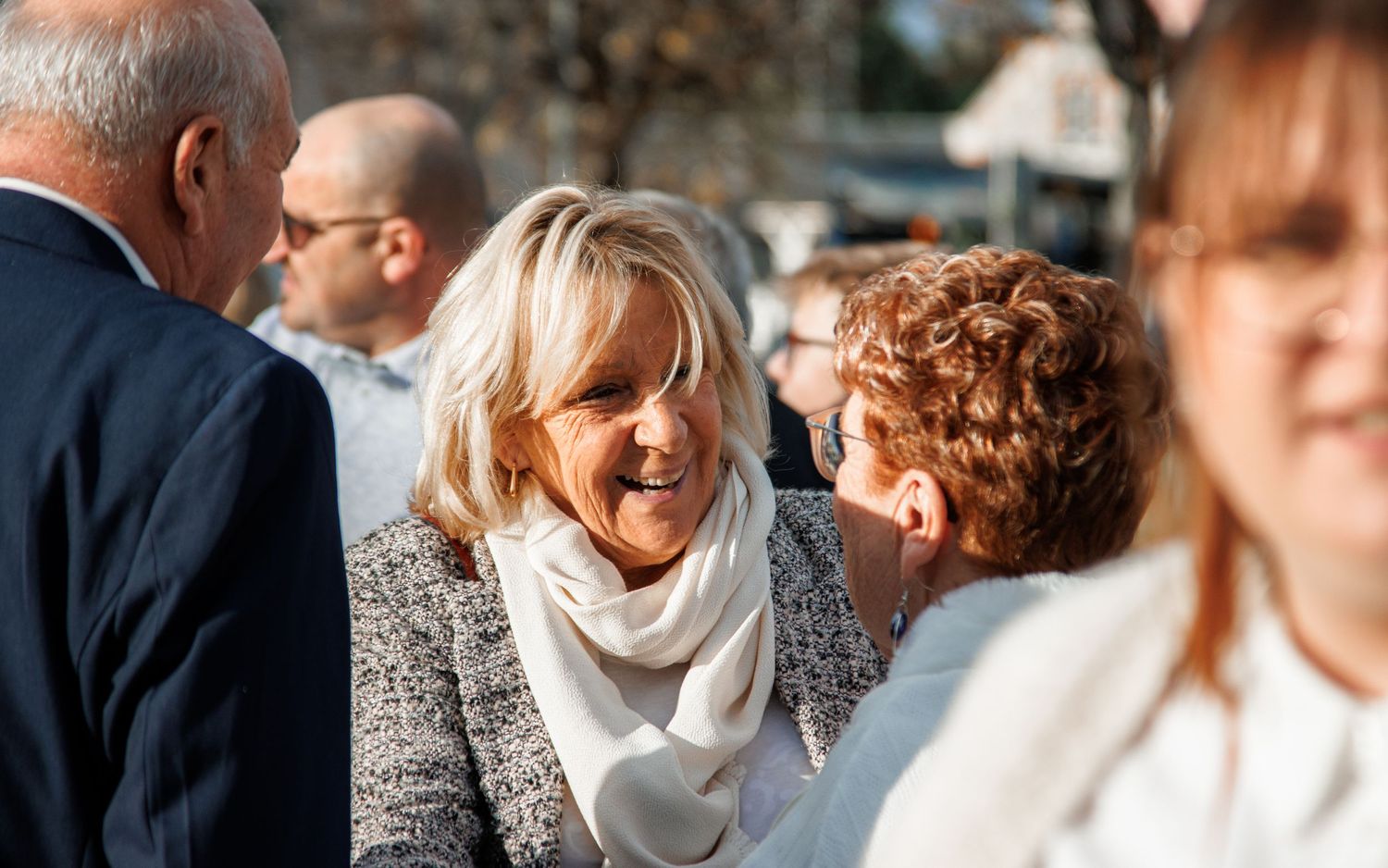 Smiling blonde woman in white scarf and gray jacket socializing outdoors at a public gathering event.