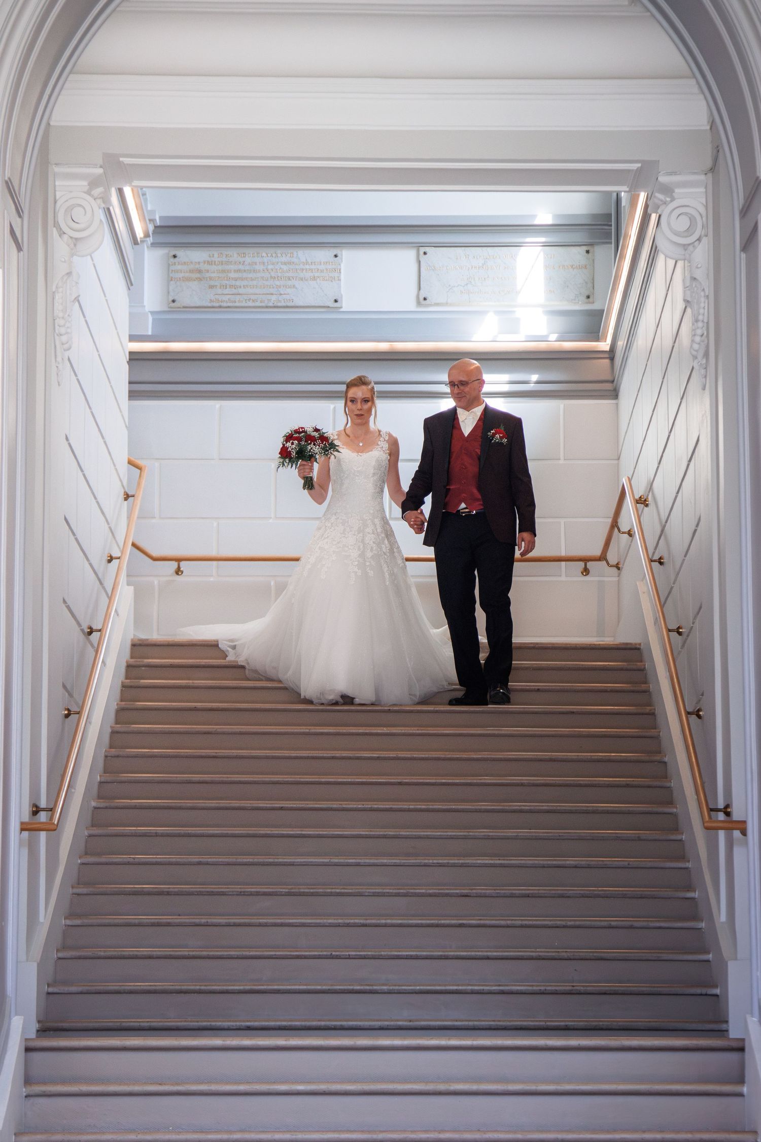 Bride in white gown and groom in dark suit descend grand staircase holding hands at wedding venue.