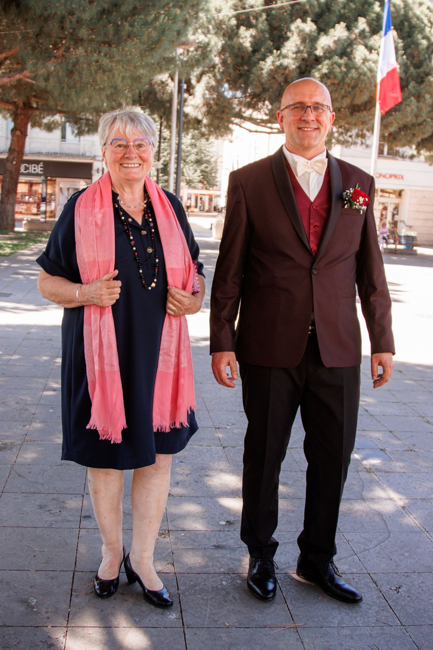A smiling couple poses outdoors near a French flag, man in burgundy suit with boutonniere.