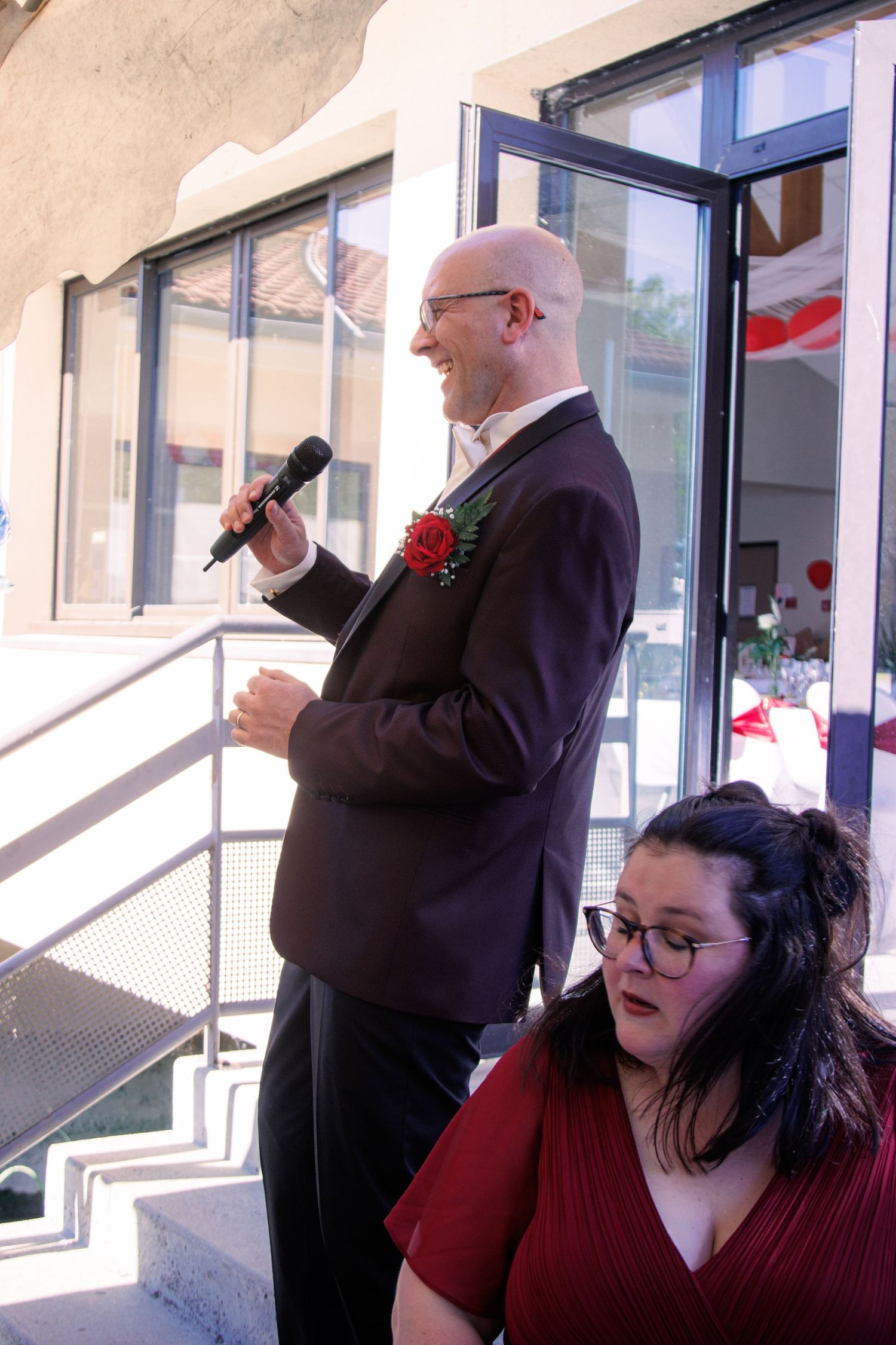 Bald groom in dark suit speaks into microphone at outdoor wedding reception while guest sits nearby.
