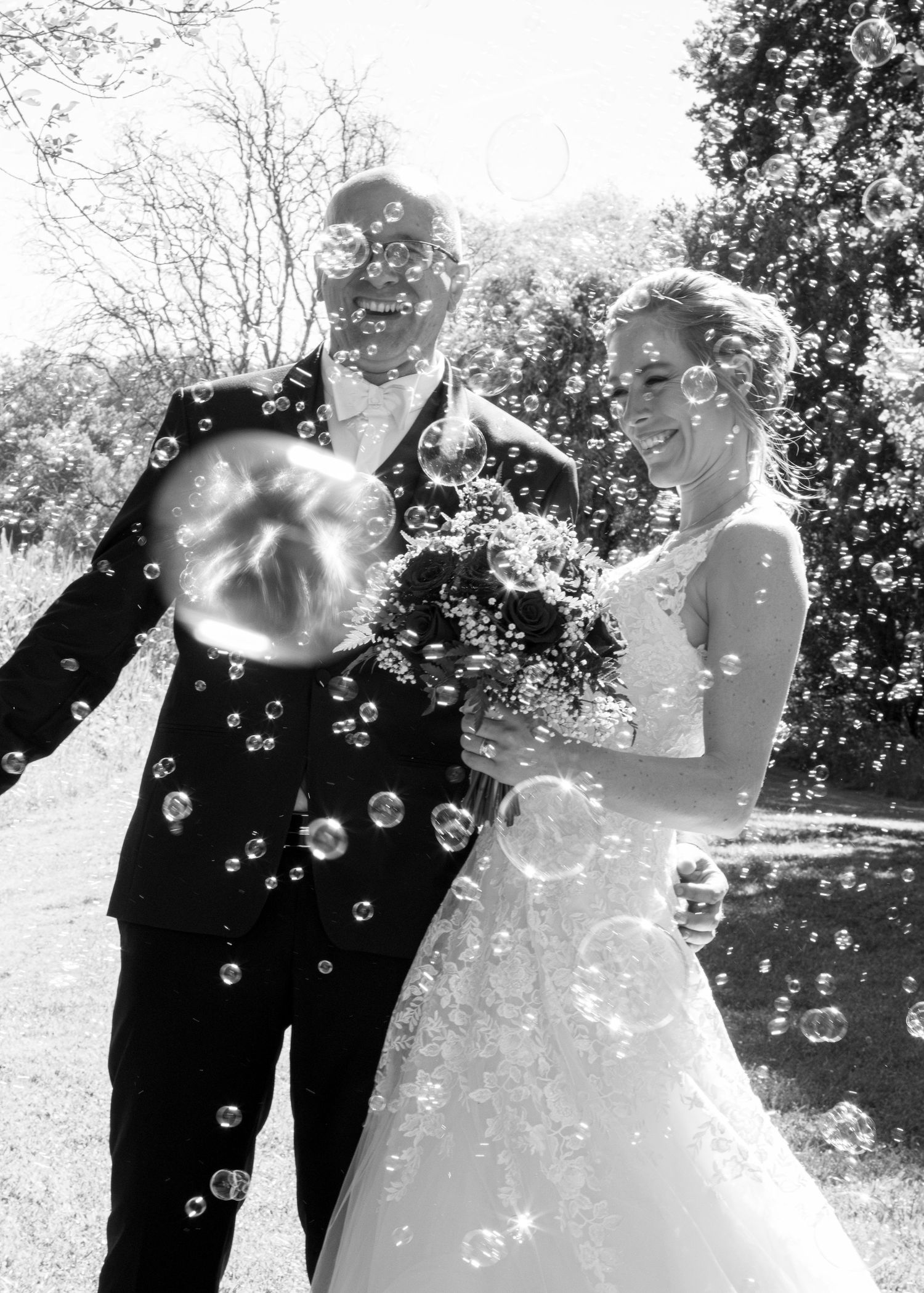 Black and white photo of laughing newlyweds surrounded by soap bubbles outdoors on their wedding day.