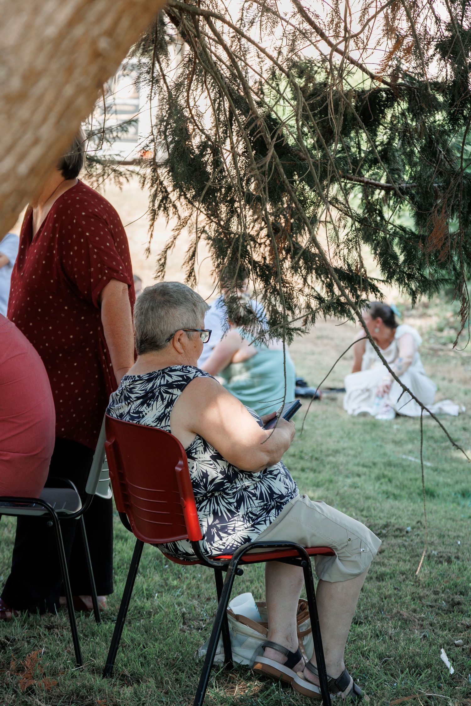 Older woman sitting on red chair using smartphone at outdoor wedding reception under tree shade.