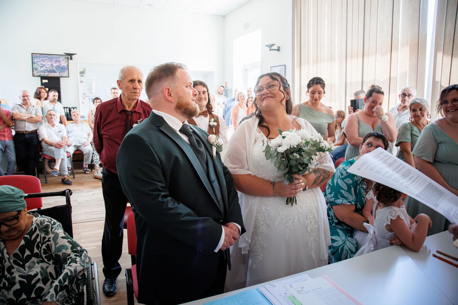 A bride and groom hold hands at their indoor wedding ceremony surrounded by seated guests in a bright venue.