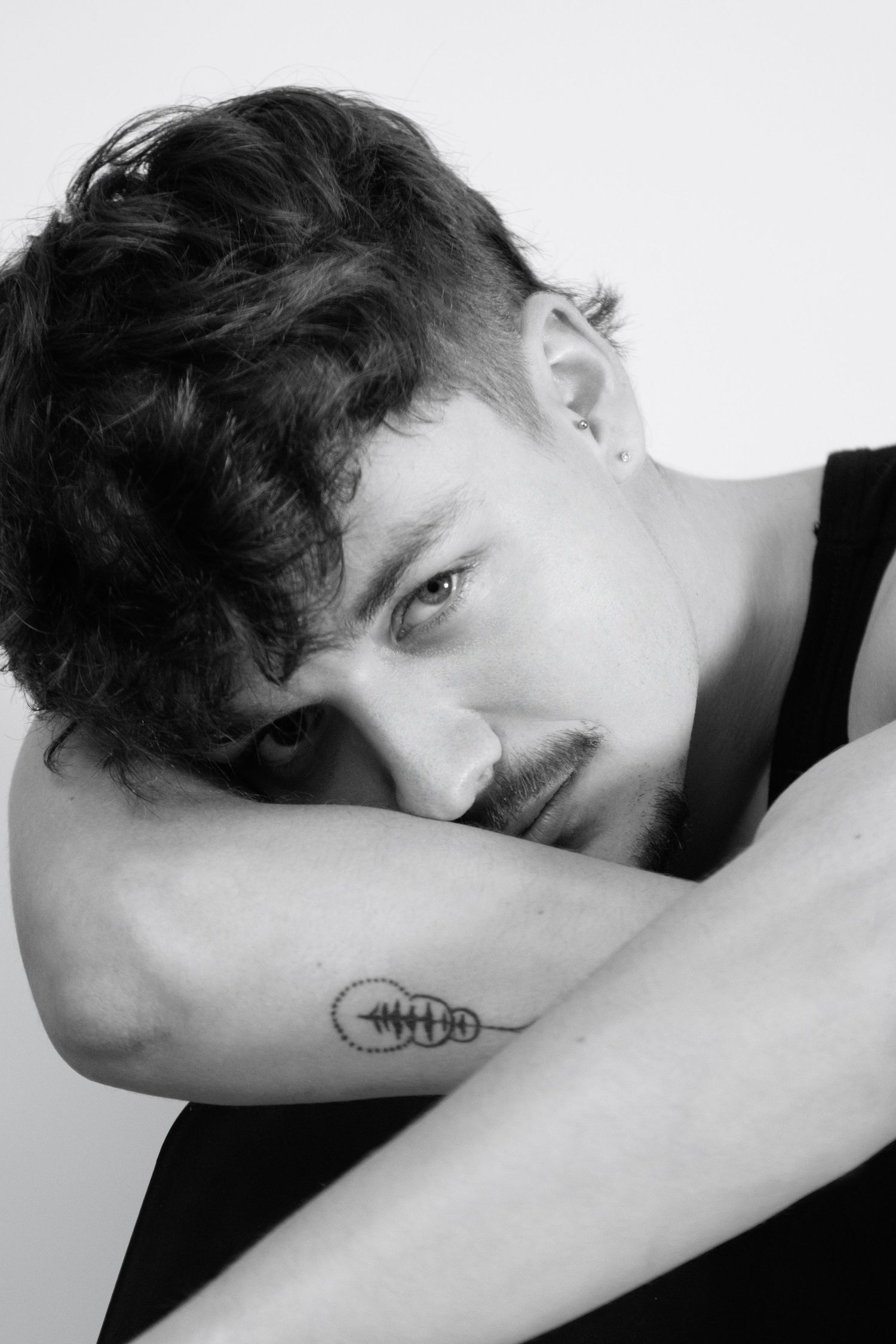 Black and white portrait of someone with dark hair wearing a black tank top and showing a small tattoo on their arm.