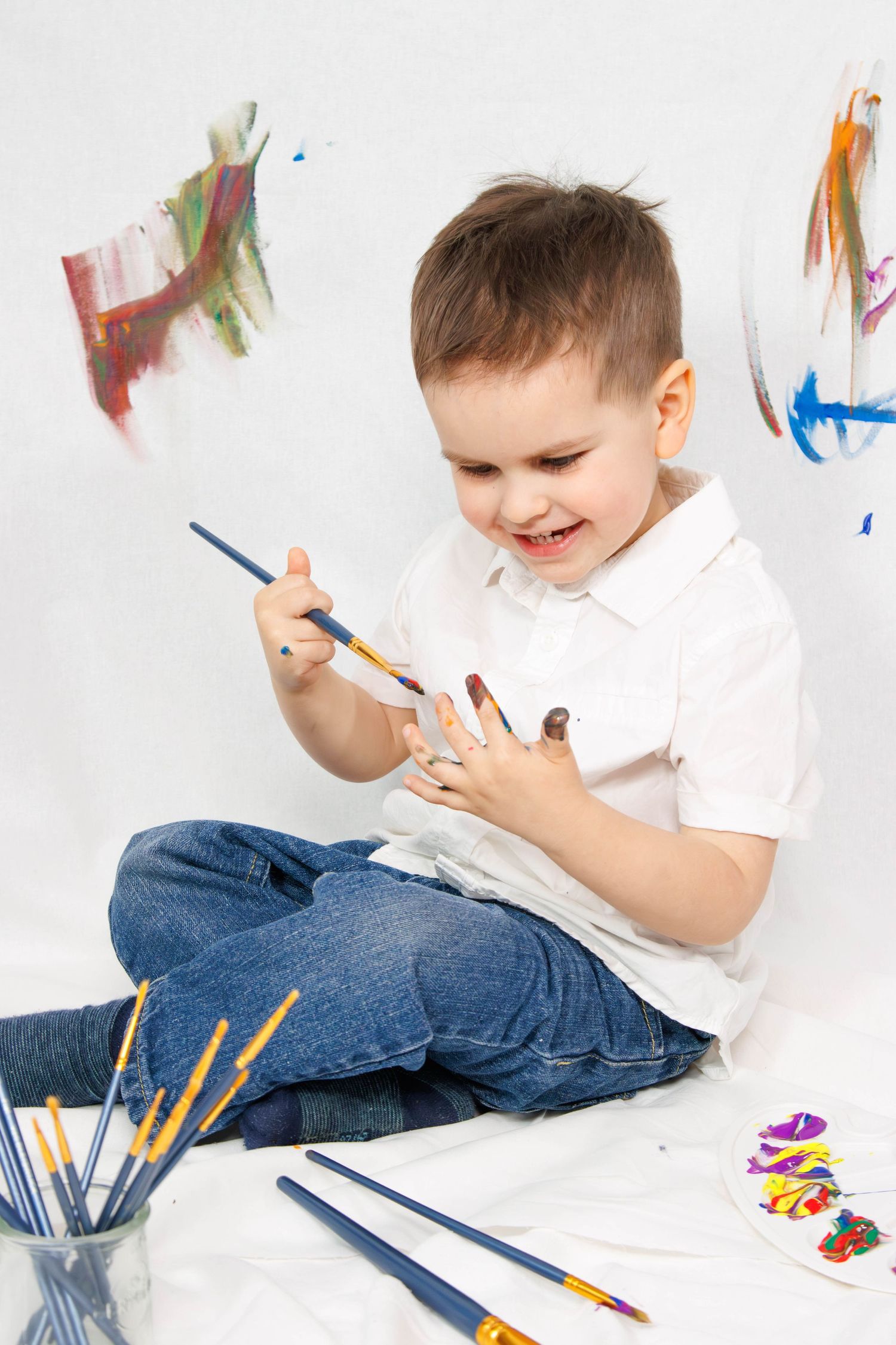 Child in white polo shirt painting while sitting cross-legged on white surface.