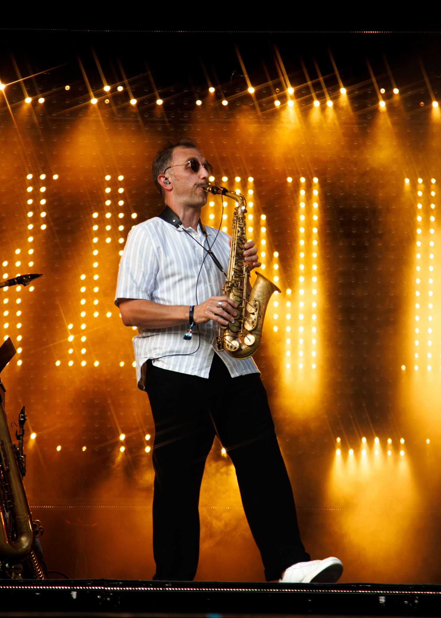 Saxophonist performing on stage under golden lights at an outdoor concert, wearing a white shirt and black pants.