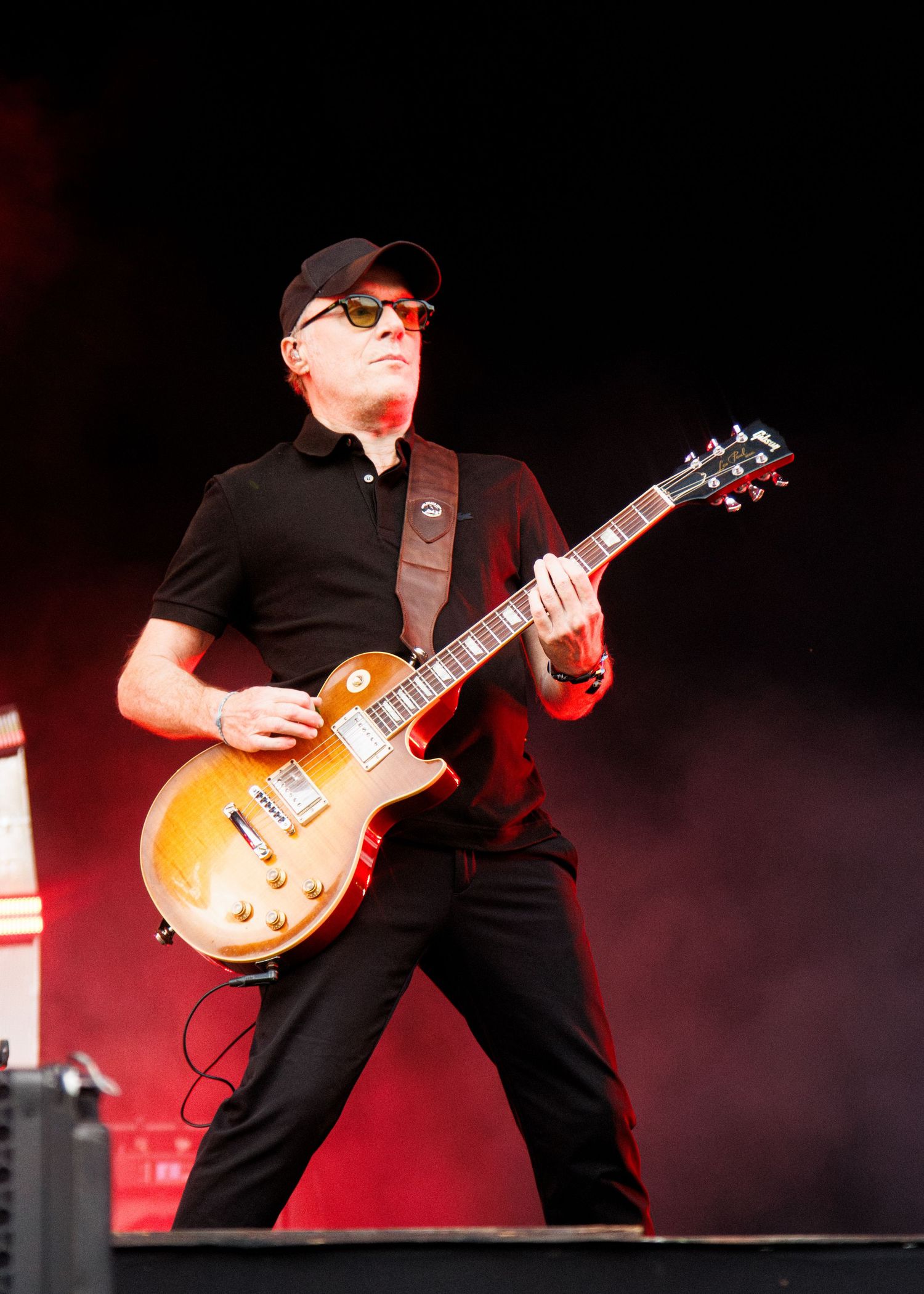 Guitarist in black outfit and cap plays a gold Les Paul electric guitar on stage under dramatic red lighting.