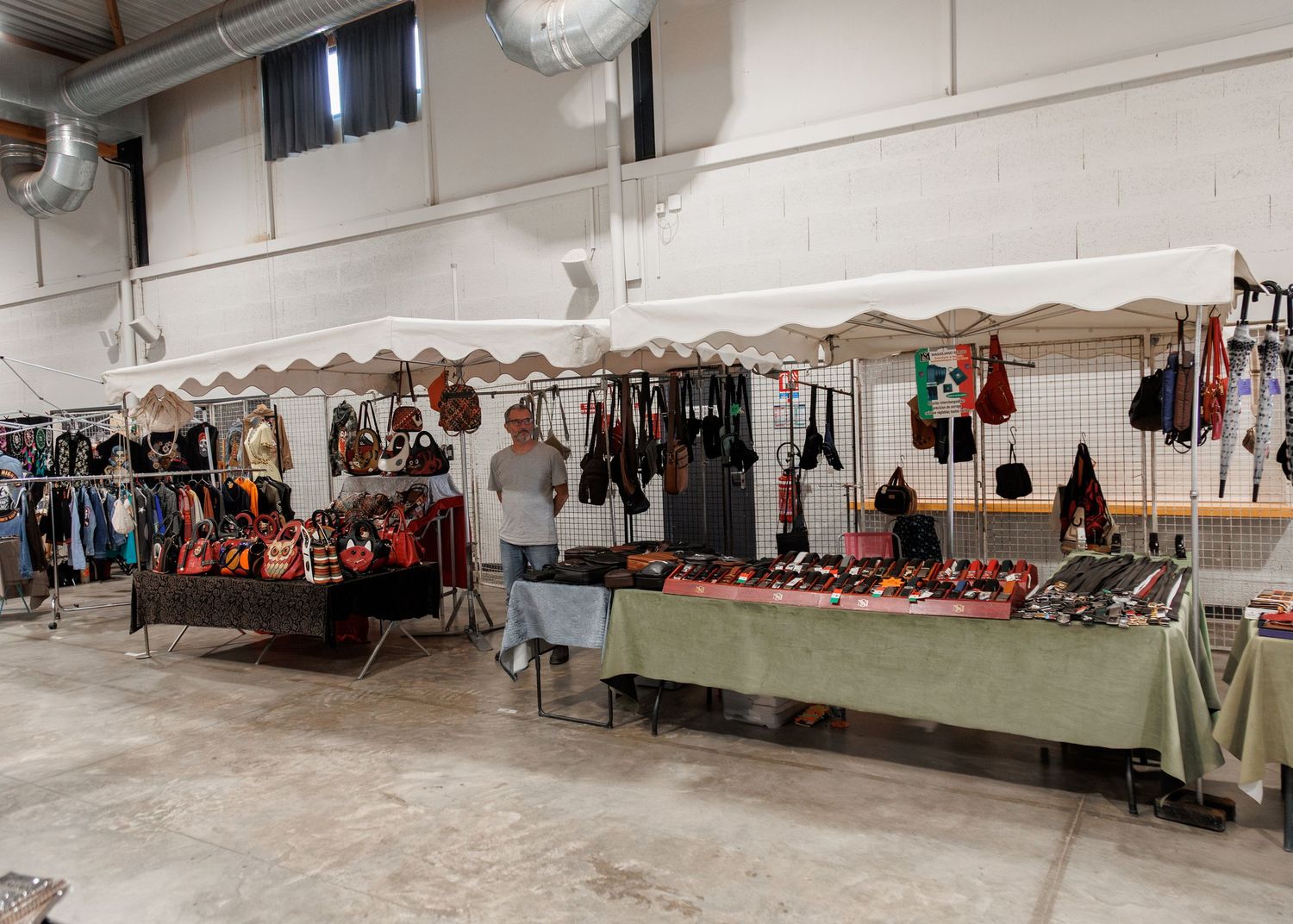 Indoor market stall displaying leather handbags, accessories, and clothing racks in a warehouse venue.