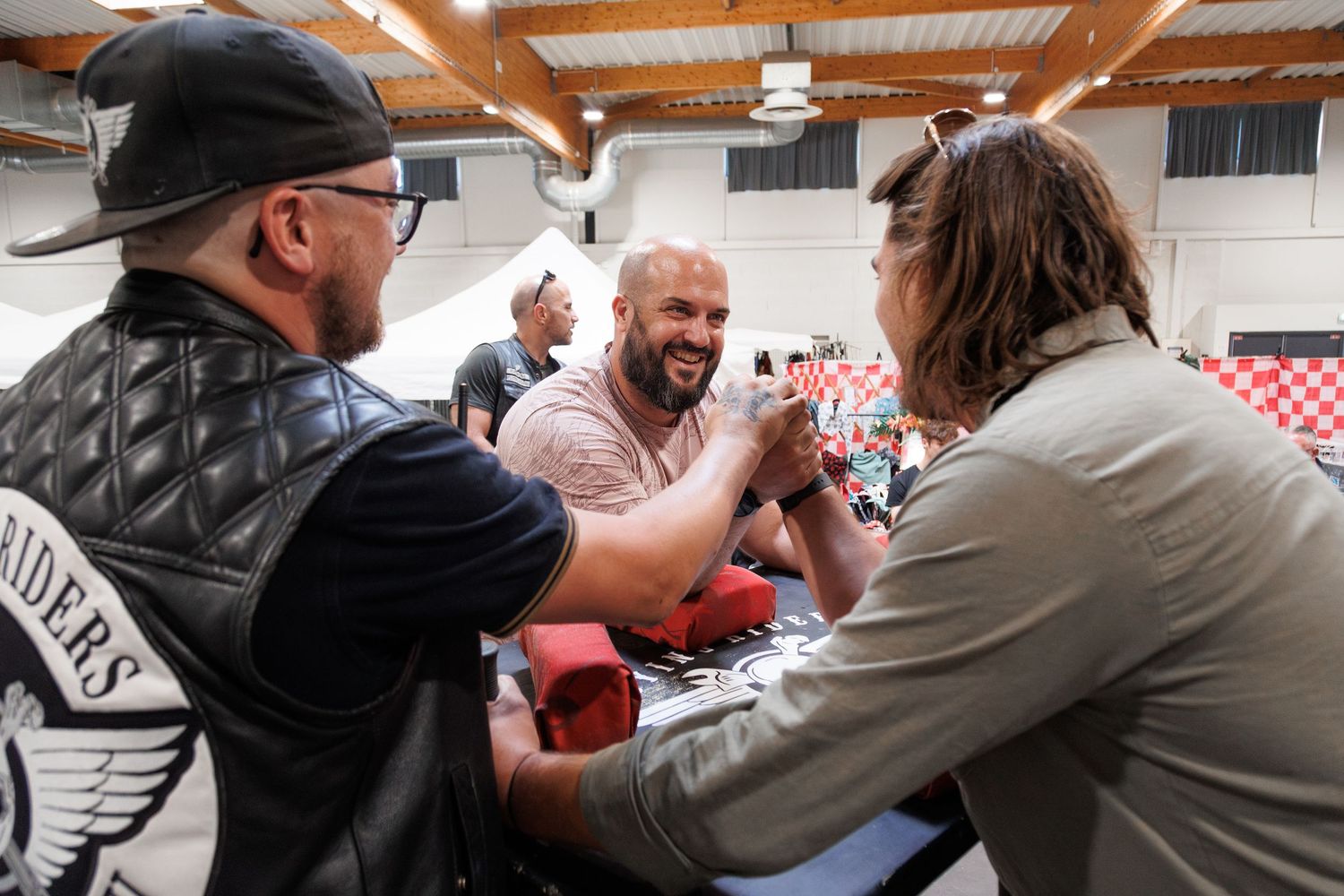 Three men arm wrestling at a table during an indoor event, smiling and engaged in friendly competition.