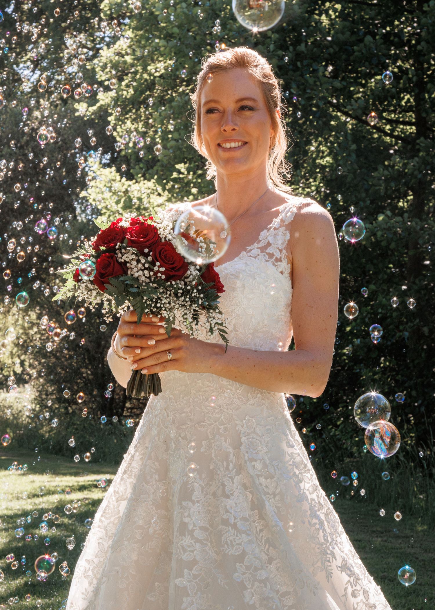 Smiling bride in lace wedding dress holds red rose bouquet surrounded by floating soap bubbles outdoors.