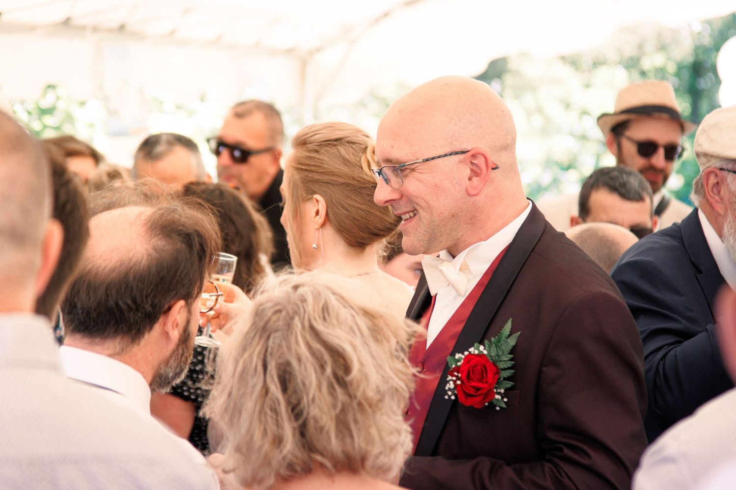 Bald groom in burgundy suit with red boutonniere smiles warmly among wedding reception guests under a tent.