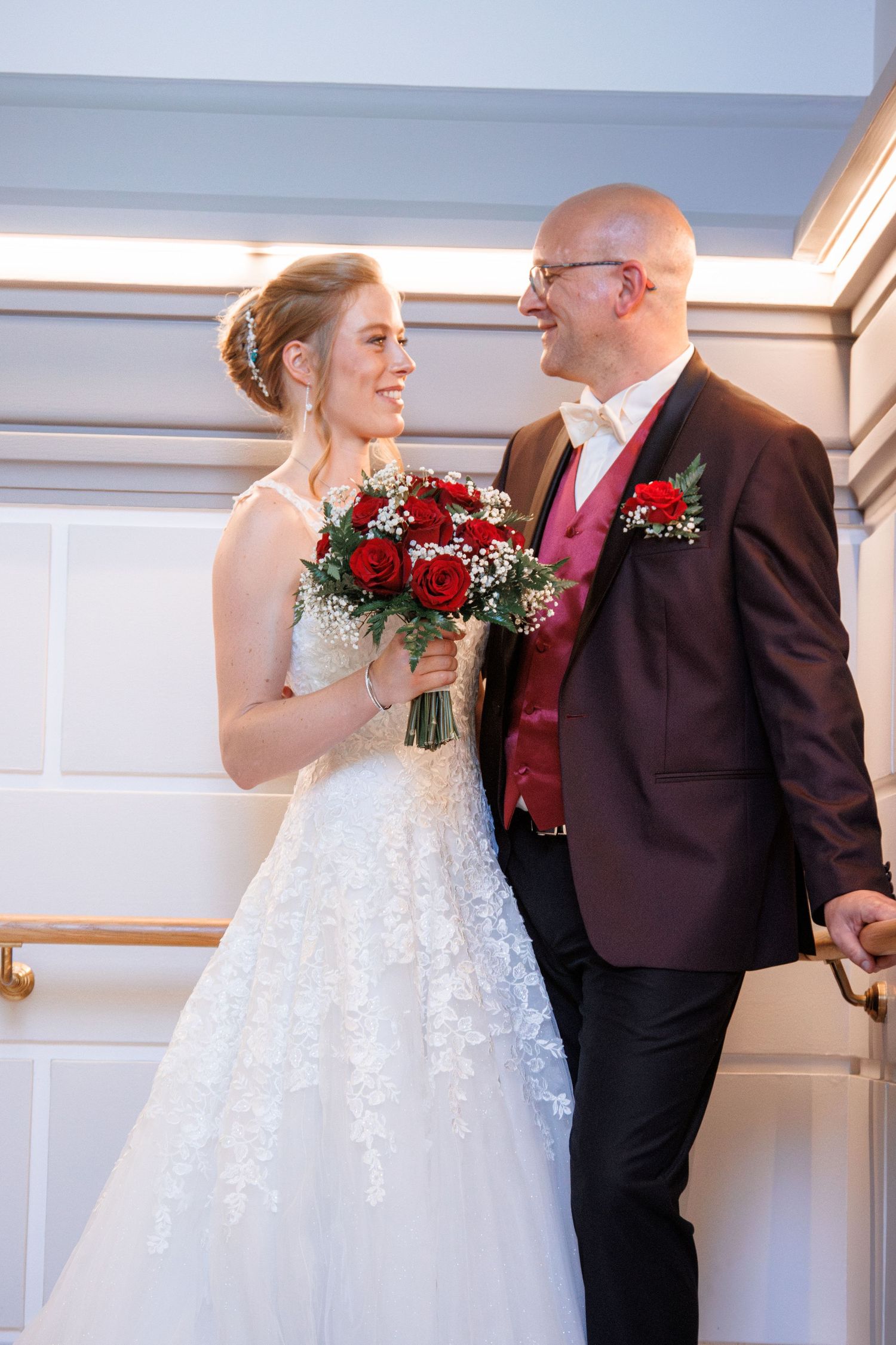 Bride in white lace gown holding red rose bouquet smiling at bald groom in burgundy suit indoors.