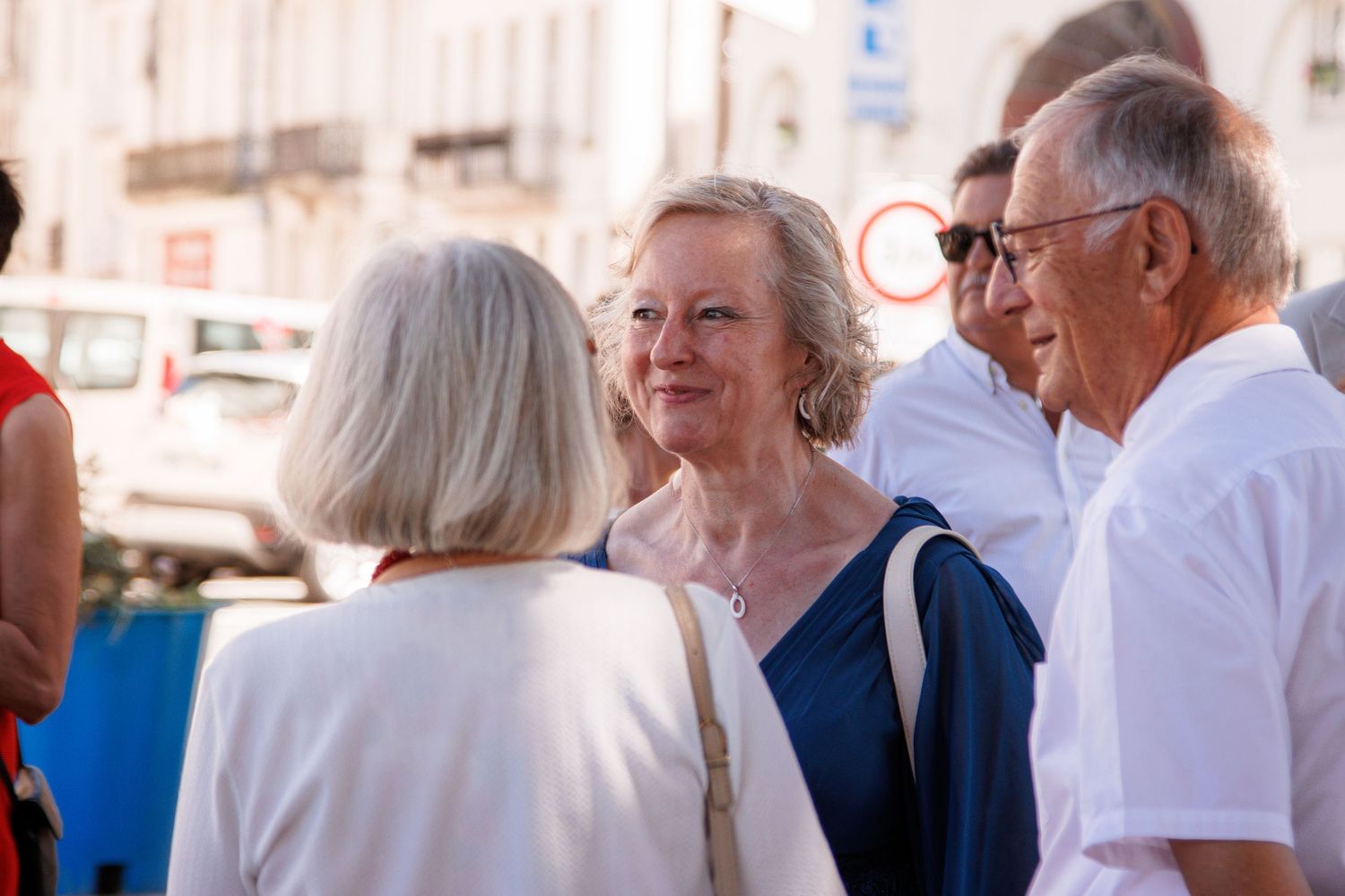 Elderly women and men socializing outdoors in a sunny European town square, dressed in summer attire.