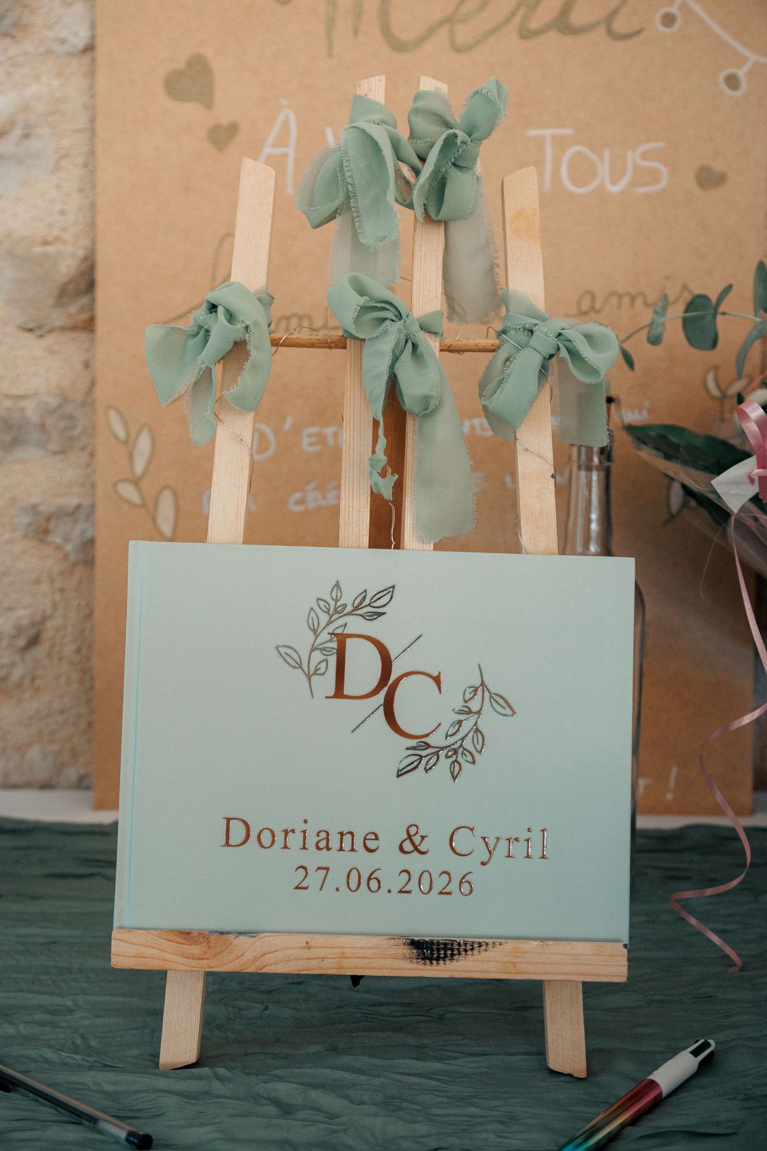 Wedding guest book sign on easel reading Doriane and Cyril 27.06.2026 with sage green ribbon bows.