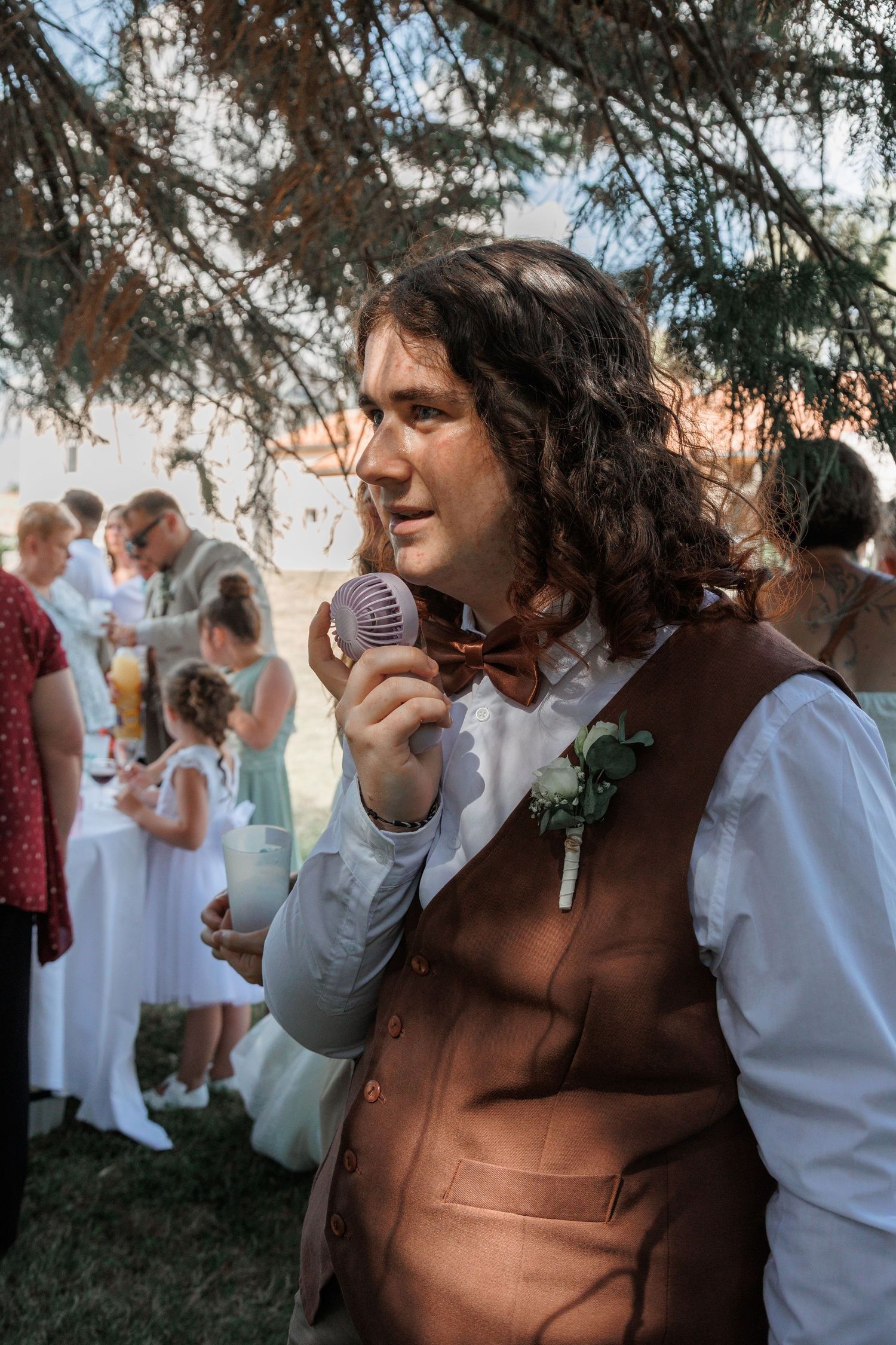 Young man with curly hair in brown vest and bow tie holds a vintage microphone at an outdoor wedding reception.