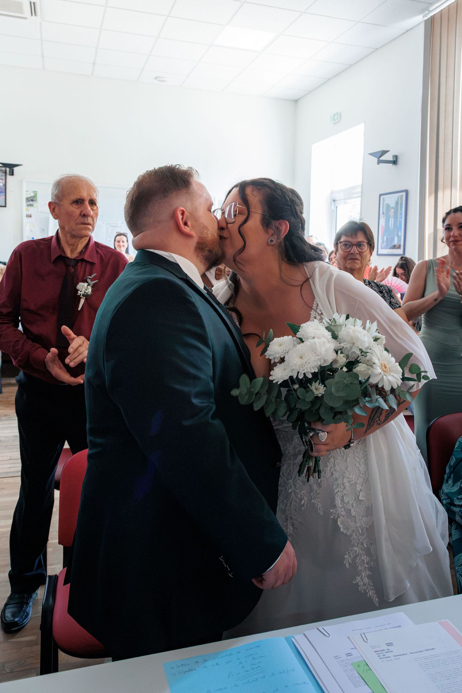 Bride and groom share their first kiss at wedding ceremony while guests applaud in background.