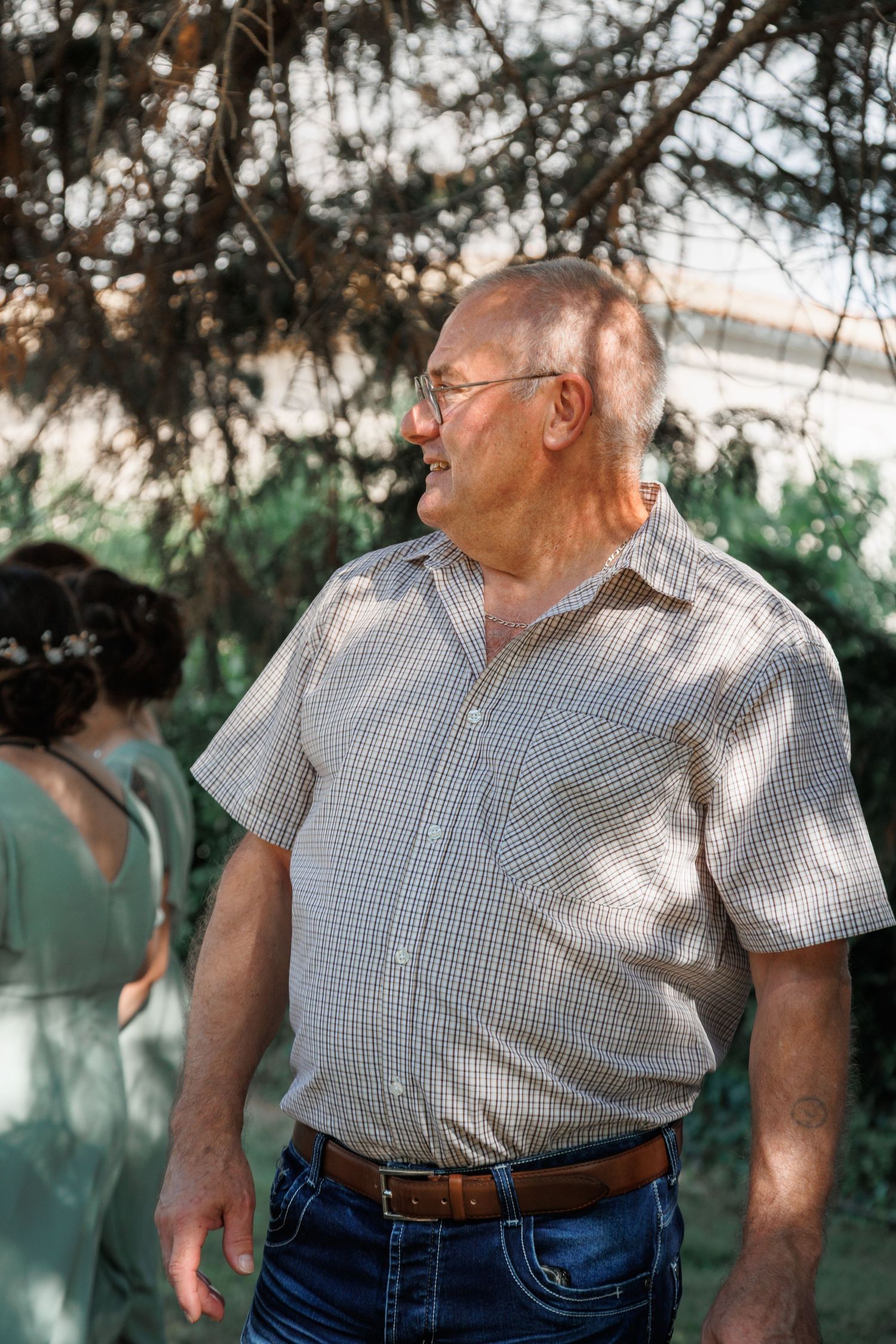 Elderly bald man with glasses smiling outdoors at a wedding reception in dappled sunlight.