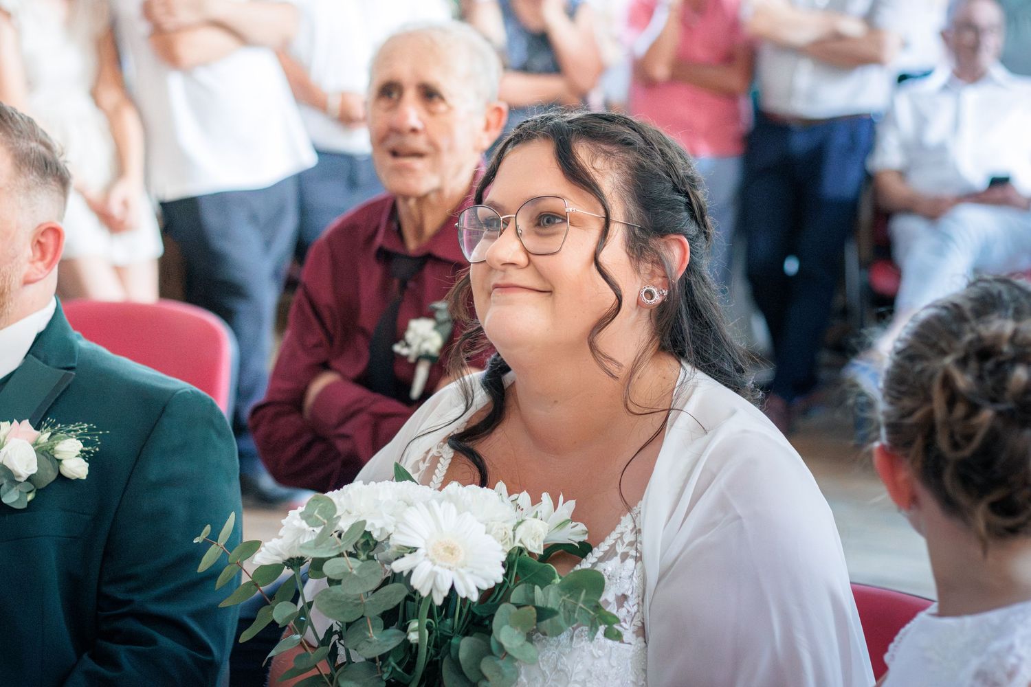 Bride holding white chrysanthemum bouquet smiling joyfully during wedding ceremony indoors.