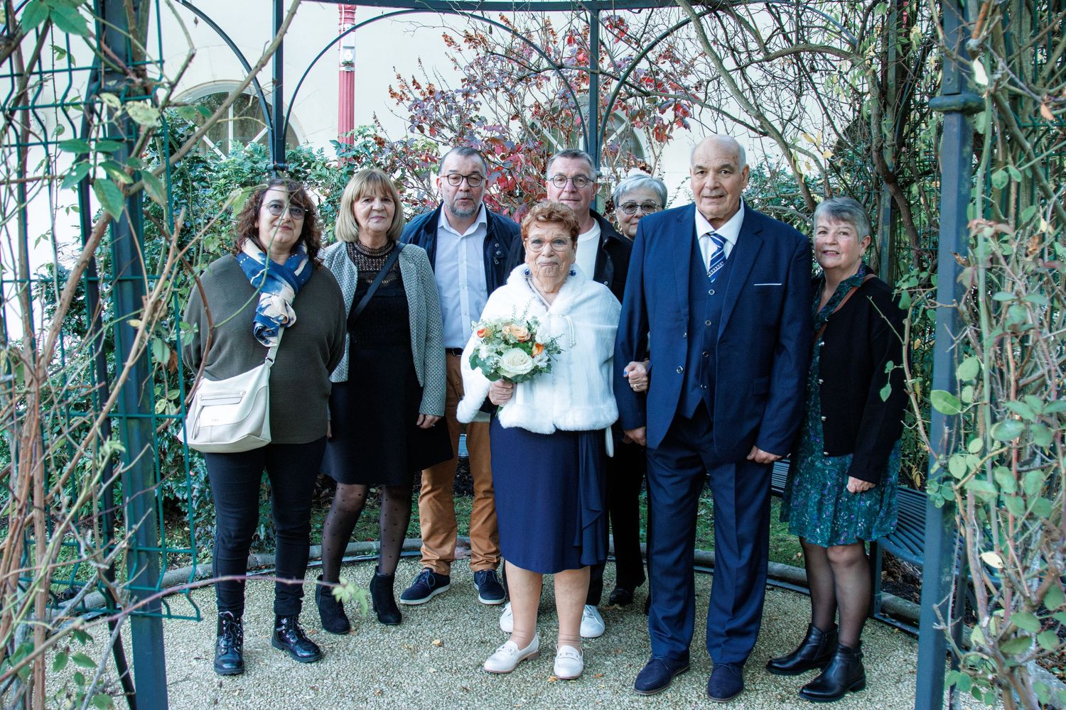 Group of people posing outdoors under a garden arch, with a woman holding a bouquet, suggesting a wedding celebration.