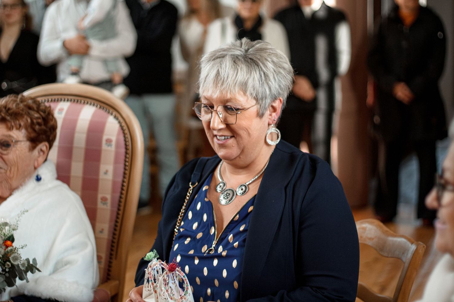 Smiling middle-aged woman with short gray hair and glasses wearing a blue polka dot dress at an elegant indoor event.